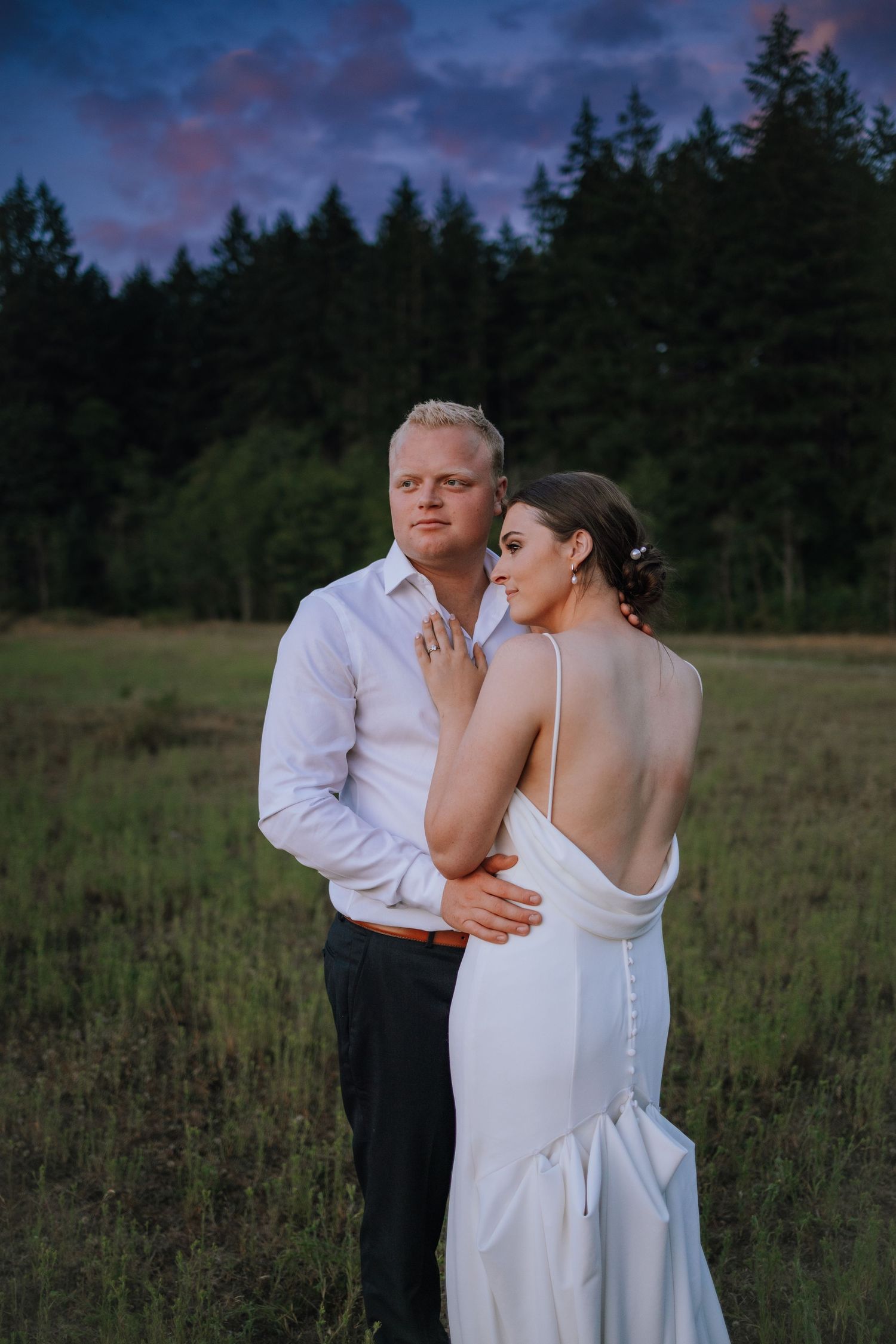 A romantic couple shares an intimate moment in a field at dusk with evergreen trees in the background.