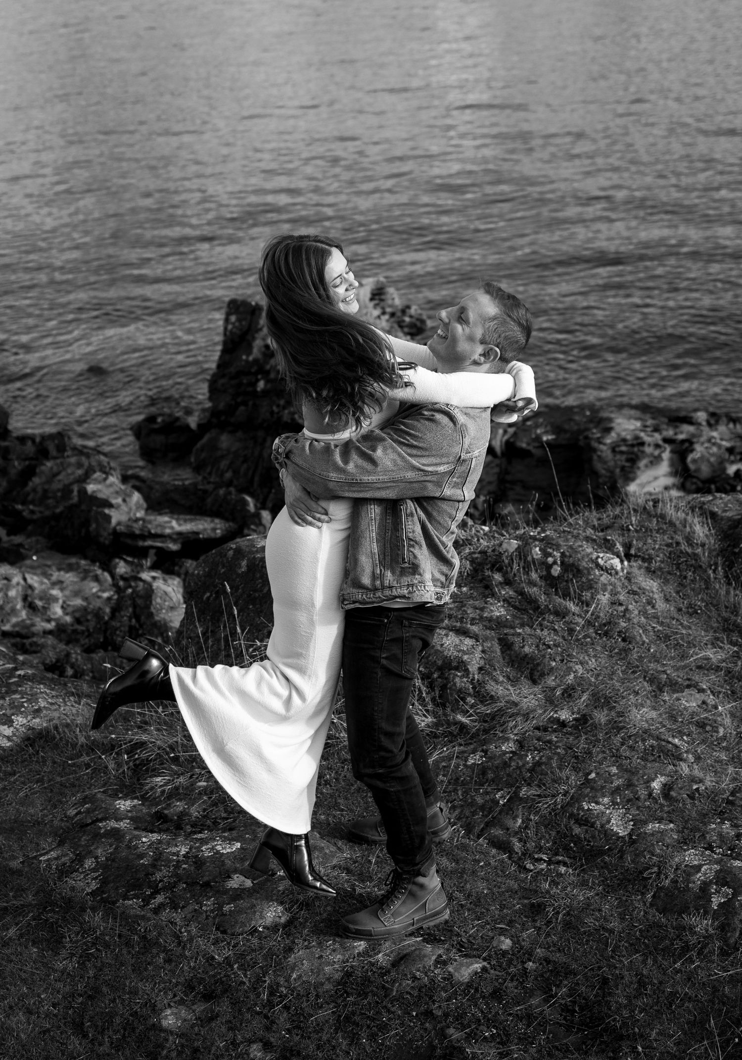 A romantic black and white photograph of a couple embracing on rocky coastal cliffs overlooking the ocean.