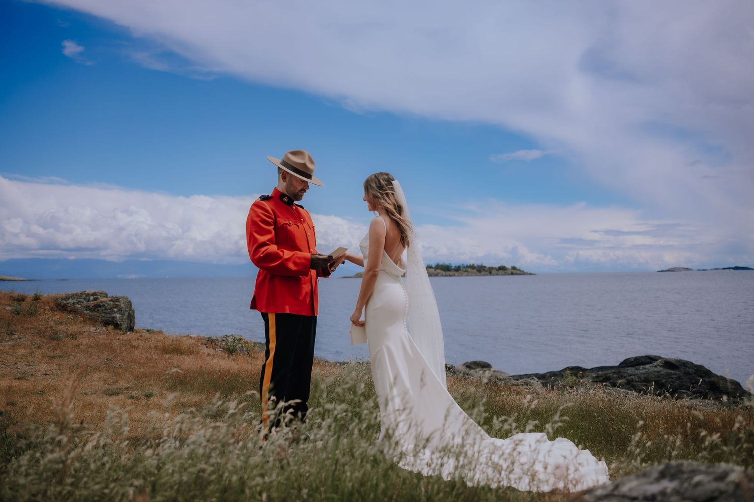 A couple poses for a wedding photo on a coastal cliff overlooking the ocean with a Canadian Mountie in uniform.