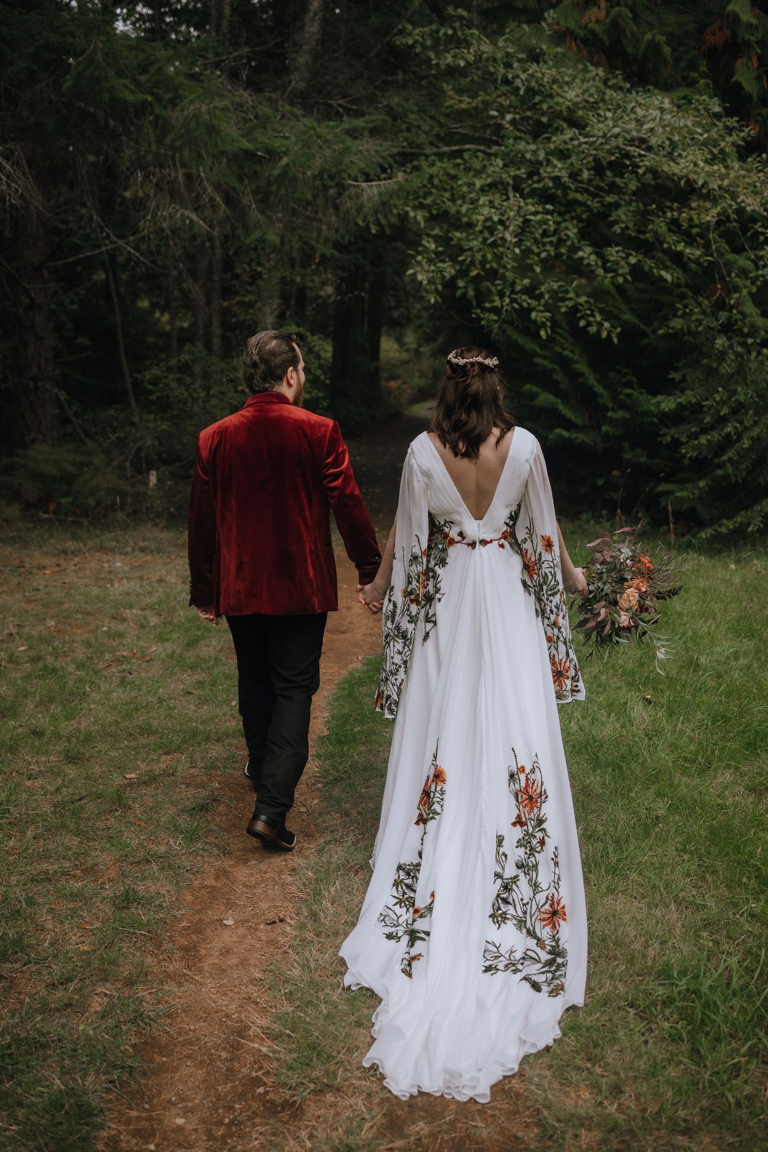 A couple walks hand in hand through a forest path, with the bride wearing an embroidered white gown and groom in red velvet.