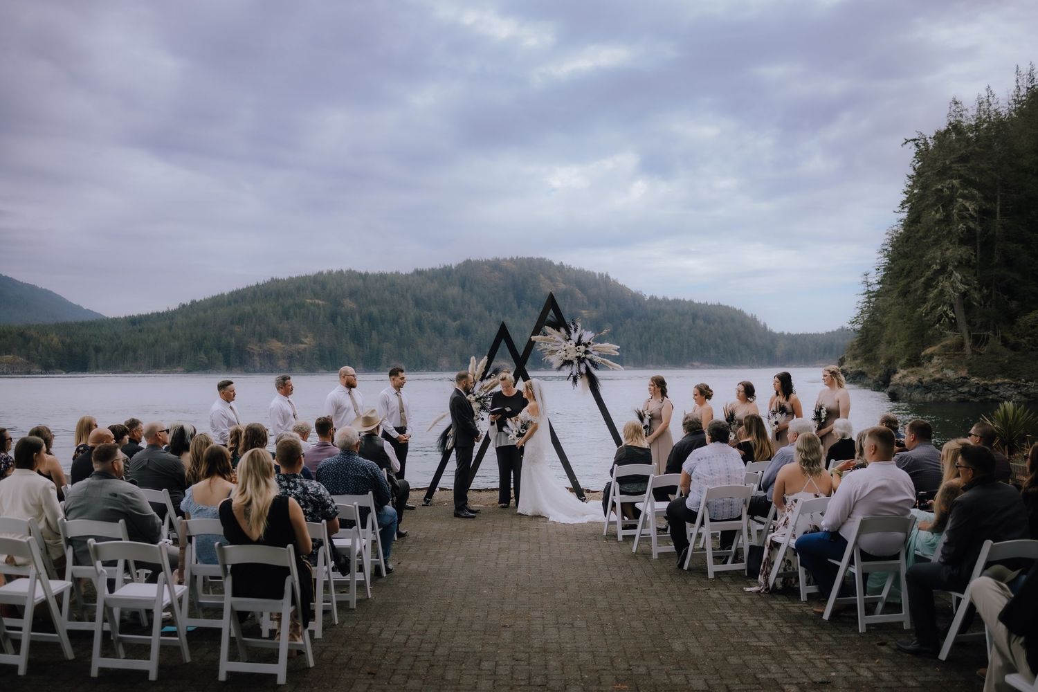 Scenic outdoor wedding ceremony setup on a misty beach with white chairs and mountains in the background.