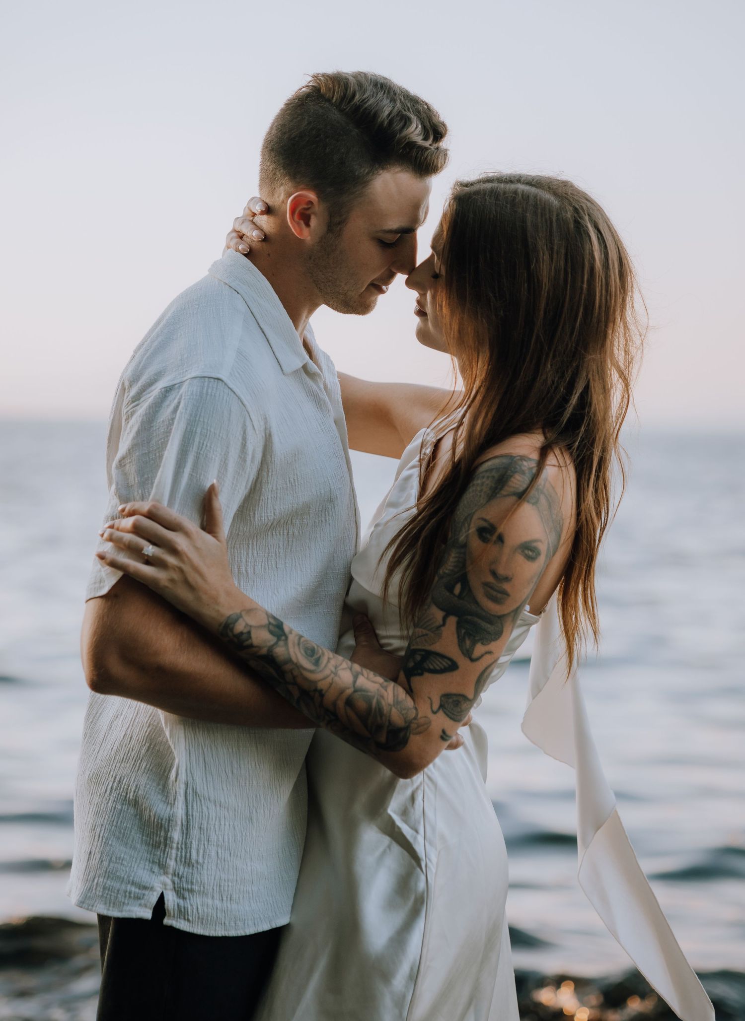 A romantic couple embraces on a misty beach at sunset in coordinating white and light colored outfits.