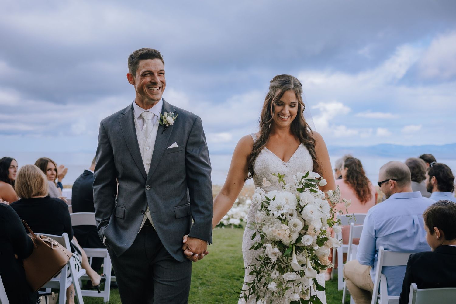 Wedding party celebrates outdoors on a grassy hillside overlooking the ocean on a partly cloudy day at The beach club resort