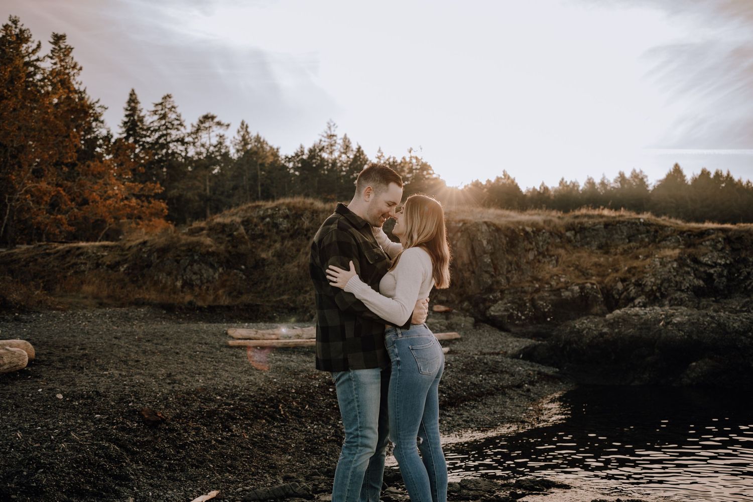 A couple embraces on a rocky cliff at sunset with autumn trees and a lake in the background.
