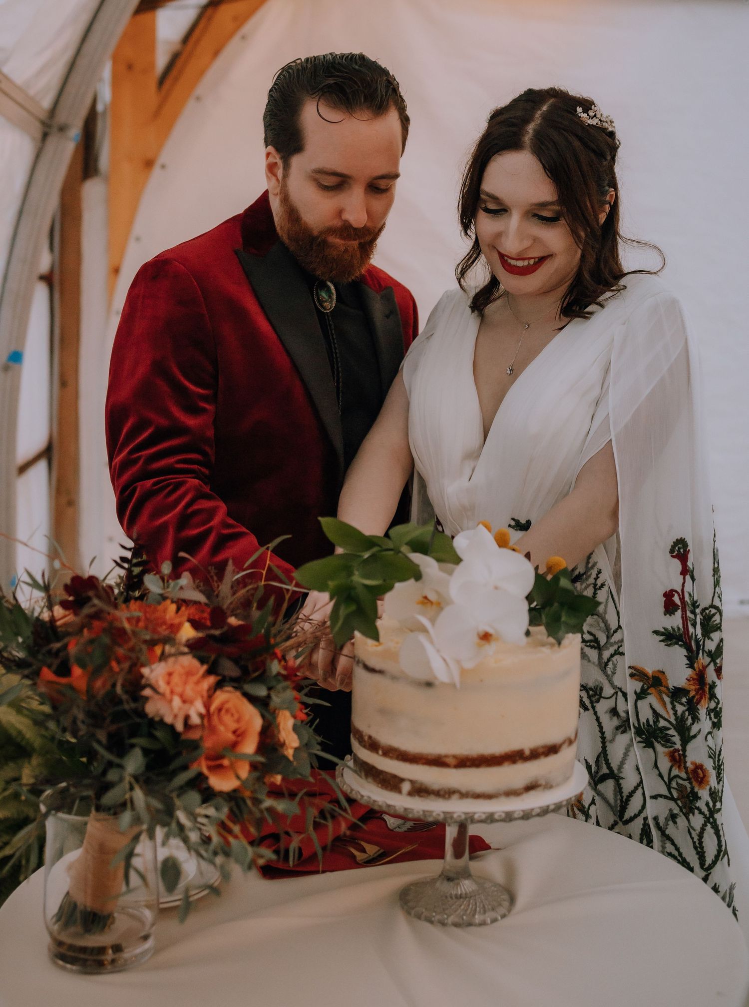 Newlywed couple cutting their elegant white wedding cake together with orange floral decor at rustic venue.