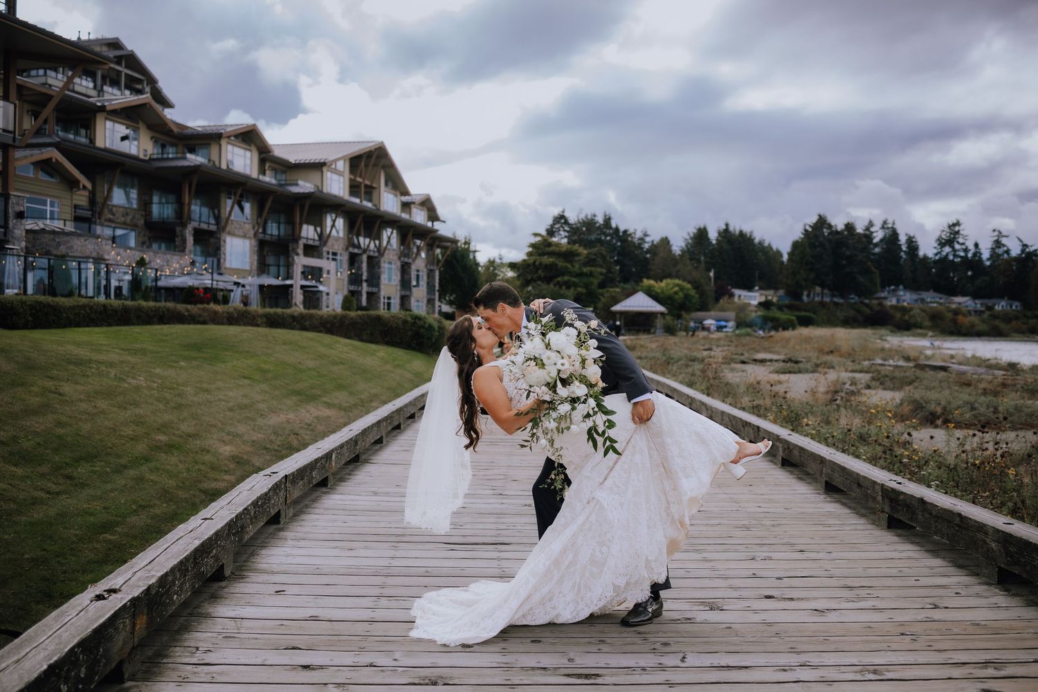 A couple in wedding attire embraces on a wooden boardwalk at Parksville beach with cloudy skies and coastal scenery in the background.