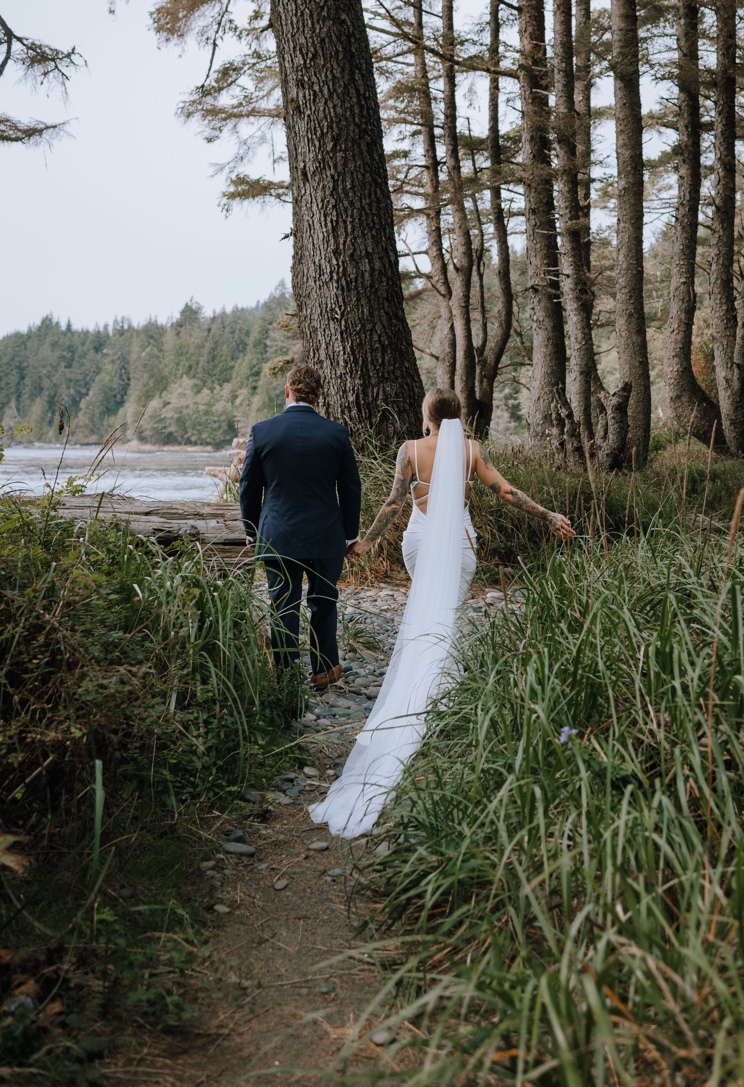 A couple walks along a scenic coastal trail through tall pine trees and grass on their wedding day.