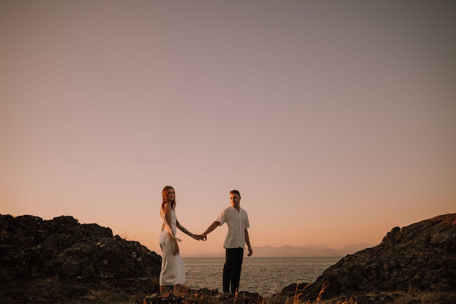 A couple holds hands while walking along a rocky beach at sunset creating silhouettes against the golden sky at sunset in Nanaimo