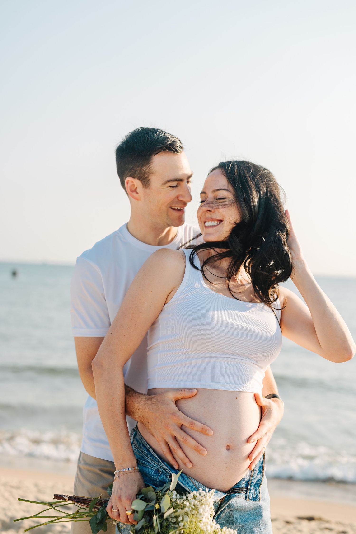 Intimate beach maternity photoshoot with baby bump and flowers at sunset.