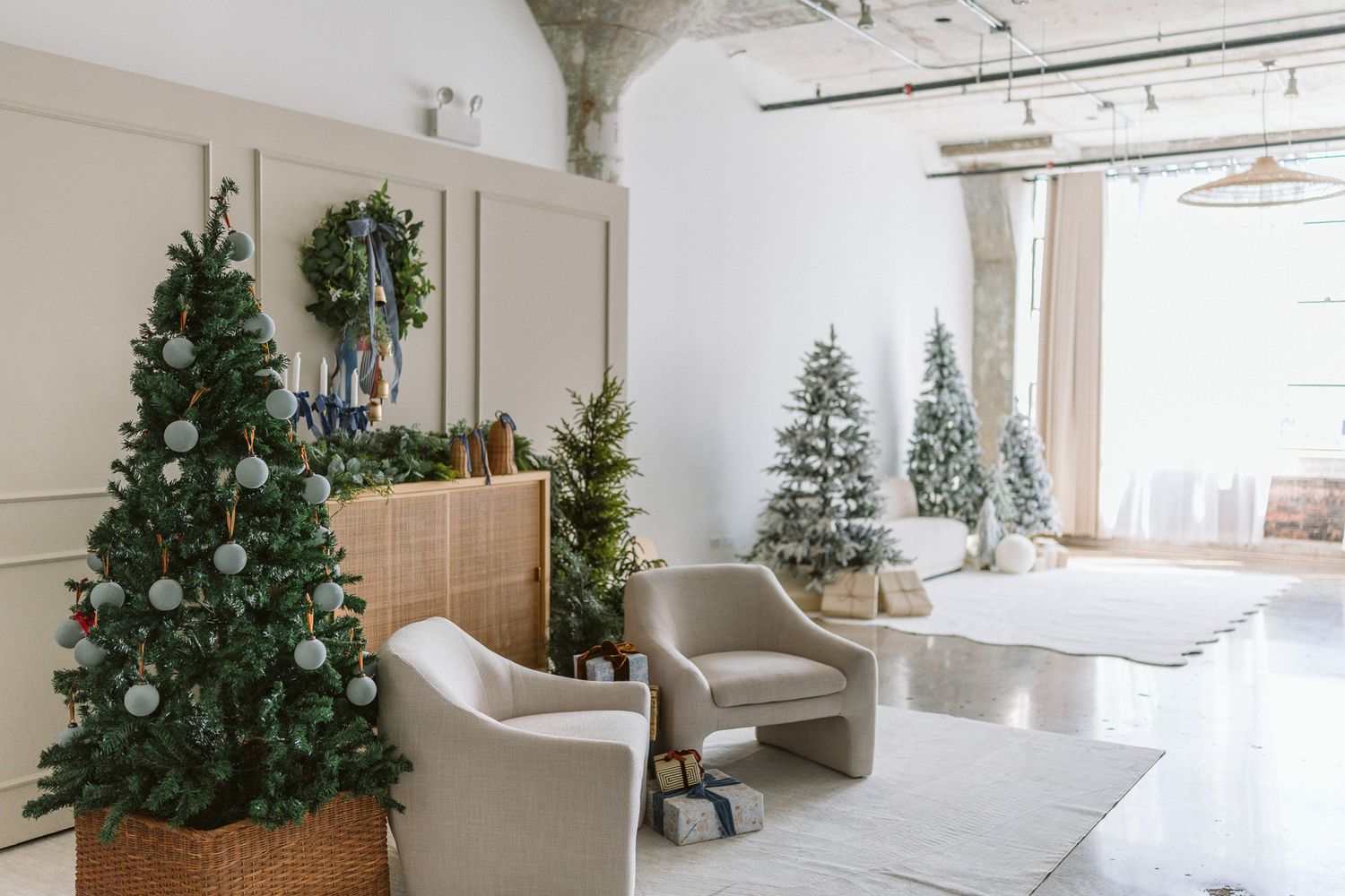 Bright loft living room decorated for Christmas with flocked trees, a wreath, cream armchairs, and wrapped gifts on a soft area rug.