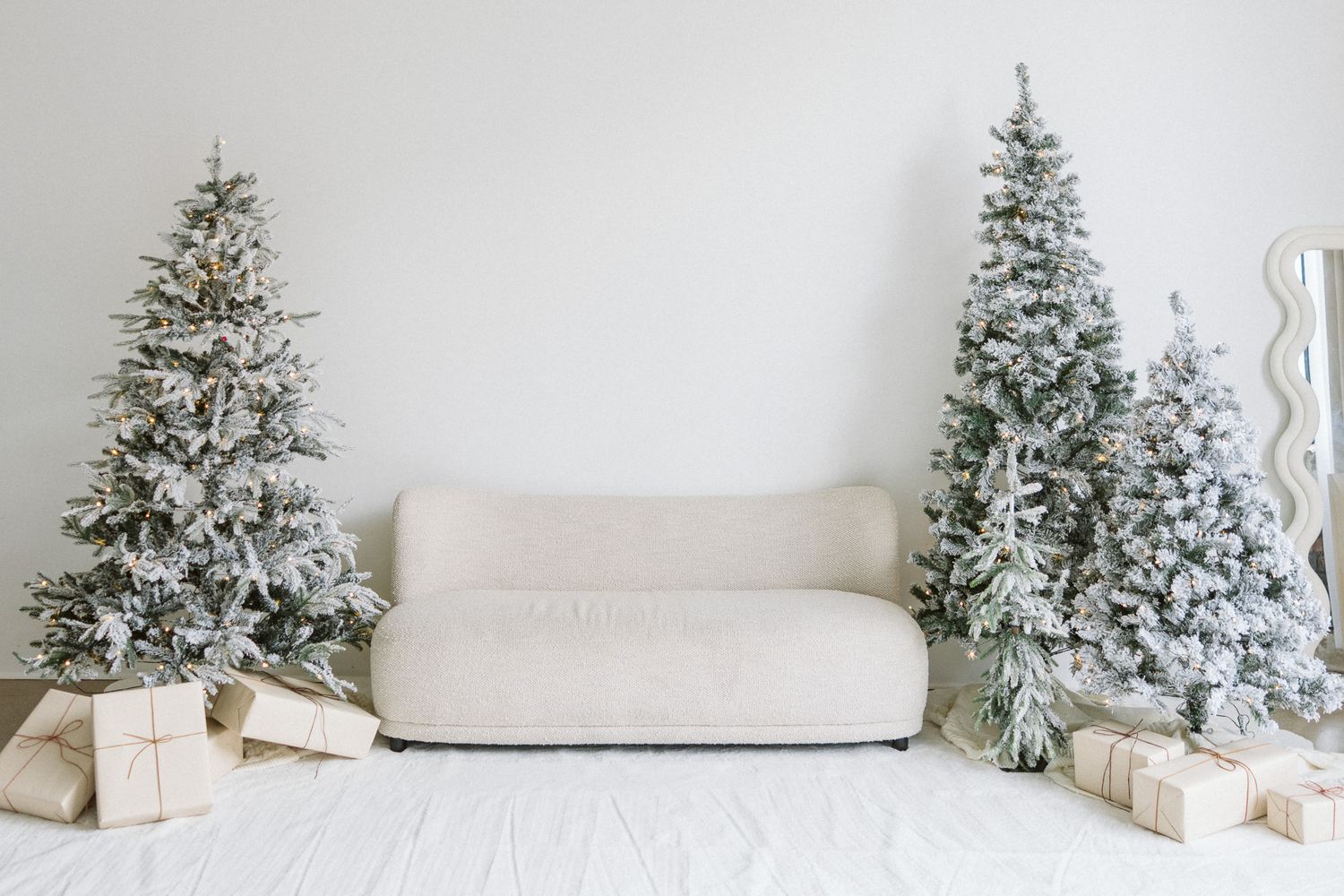 Minimalist white living room with a cream boucle sofa between flocked snowy Christmas trees and wrapped gift boxes on a soft white rug.