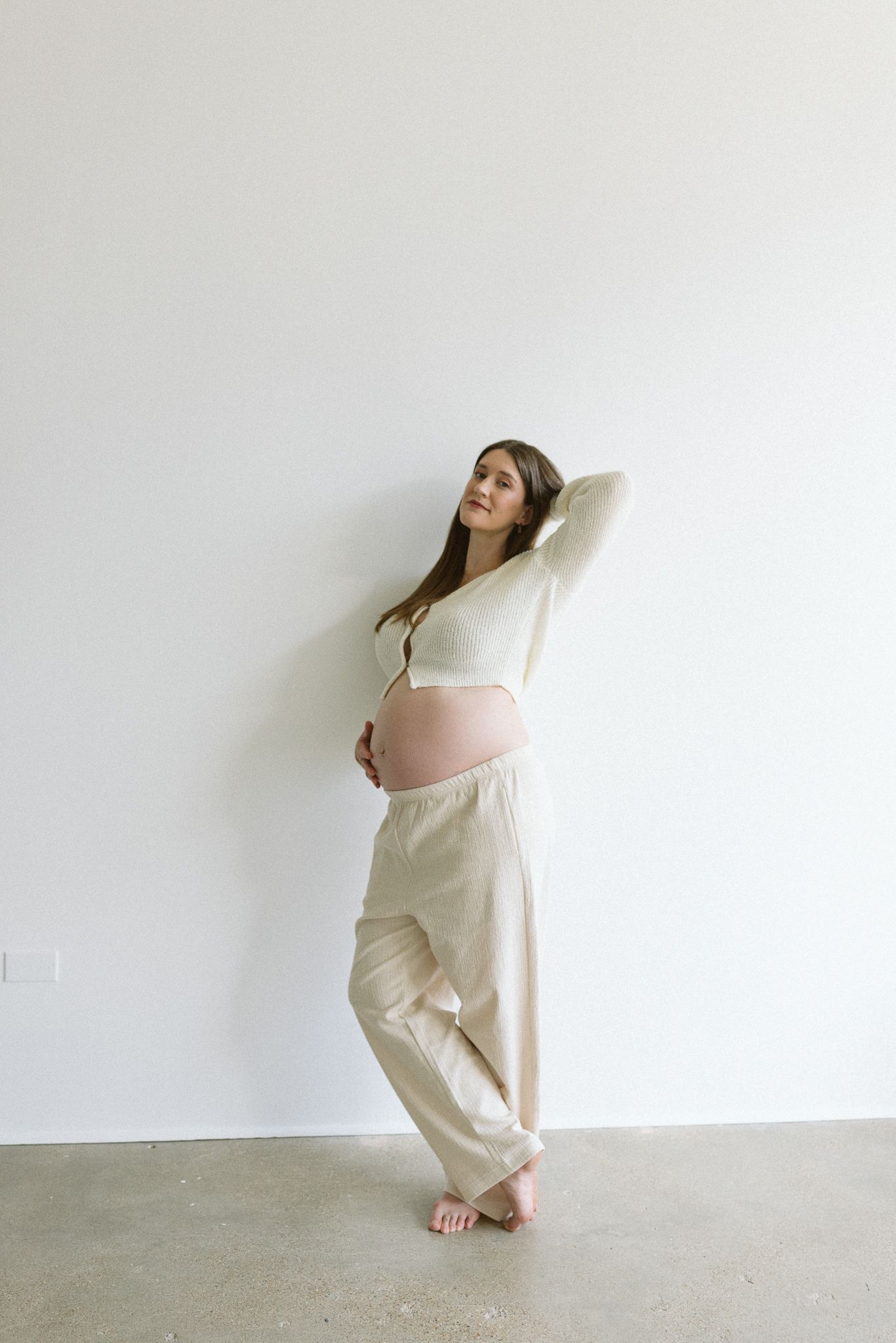 Woman in cream-colored loungewear stands against white wall with relaxed pose and bare feet.