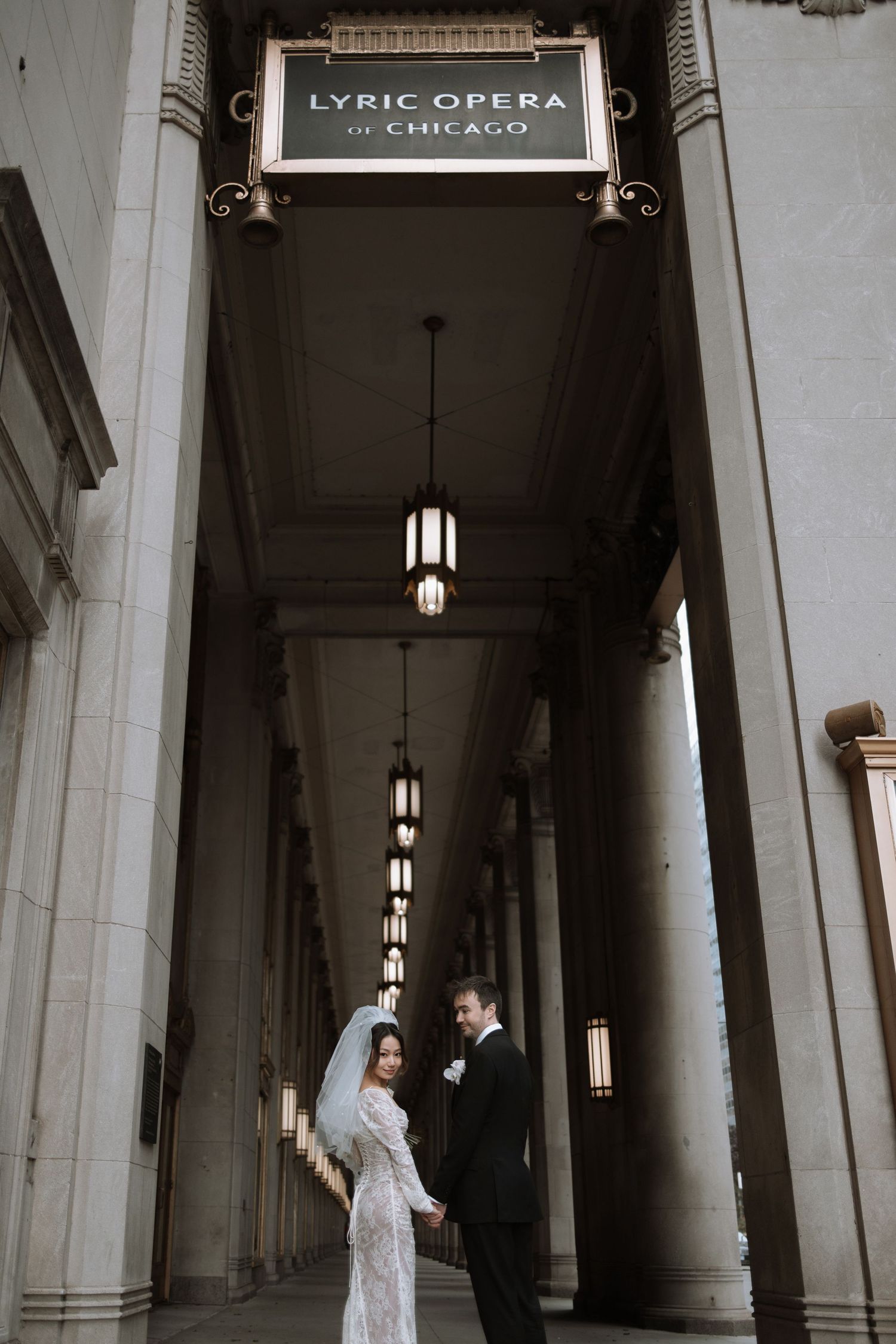 A couple stands in a dramatic colonnade beneath the historic Lyric Opera of Chicago sign with hanging pendant lights.