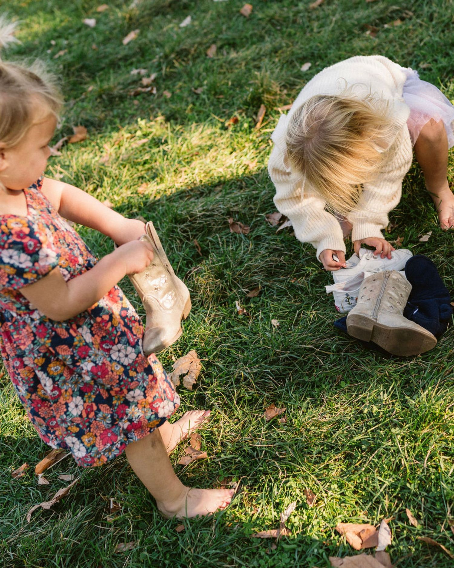Two young children playing on green grass, one in a floral dress, the other in a cream sweater bending down.