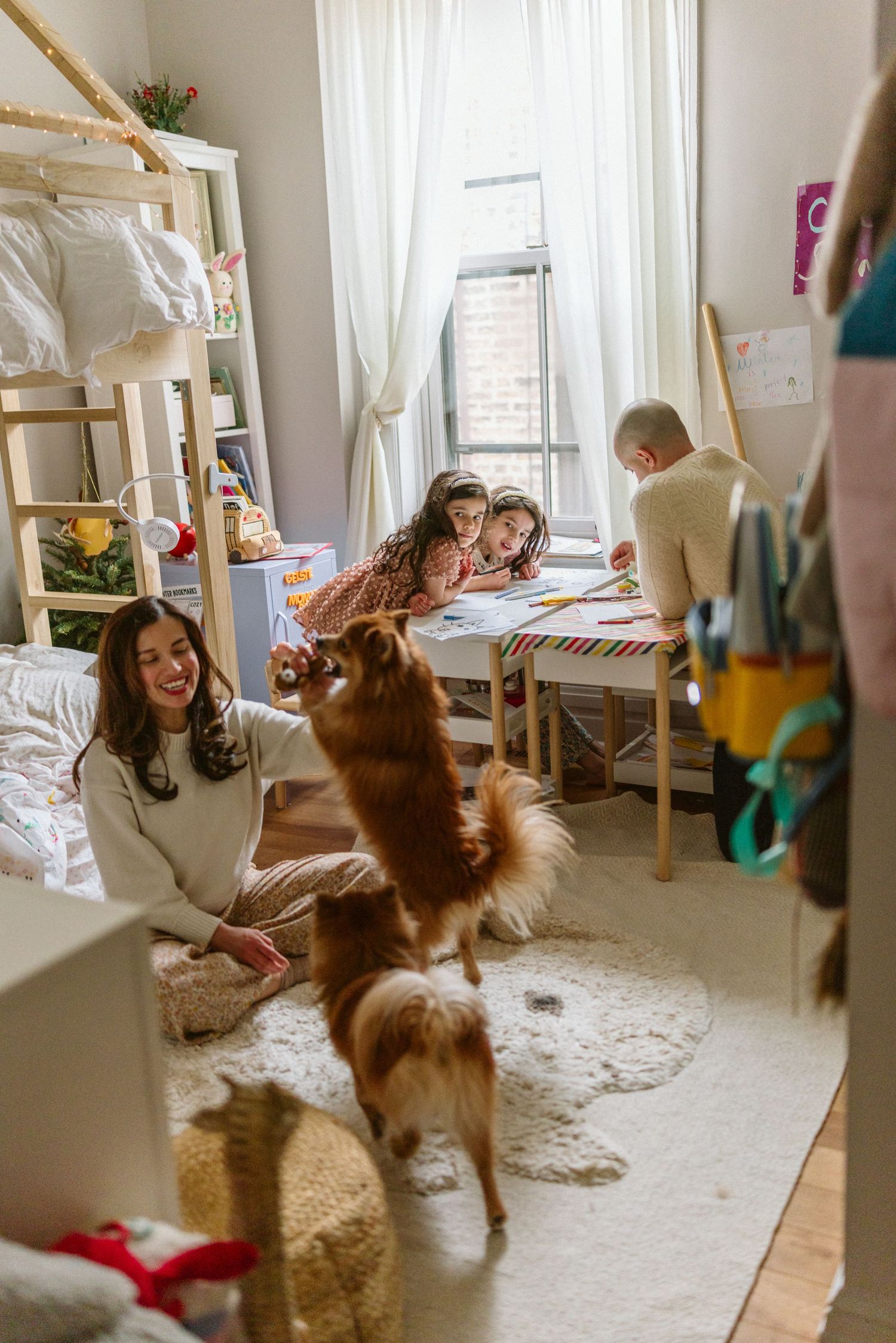 Family enjoying time in a cozy kids bedroom with a playful brown dog, children drawing at a desk while mom sits on the floor.