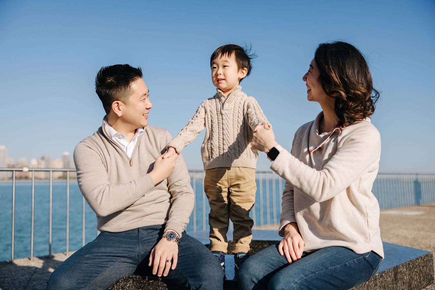 A happy Asian family with two parents and a young toddler sitting on a stone wall by the waterfront on a sunny day.