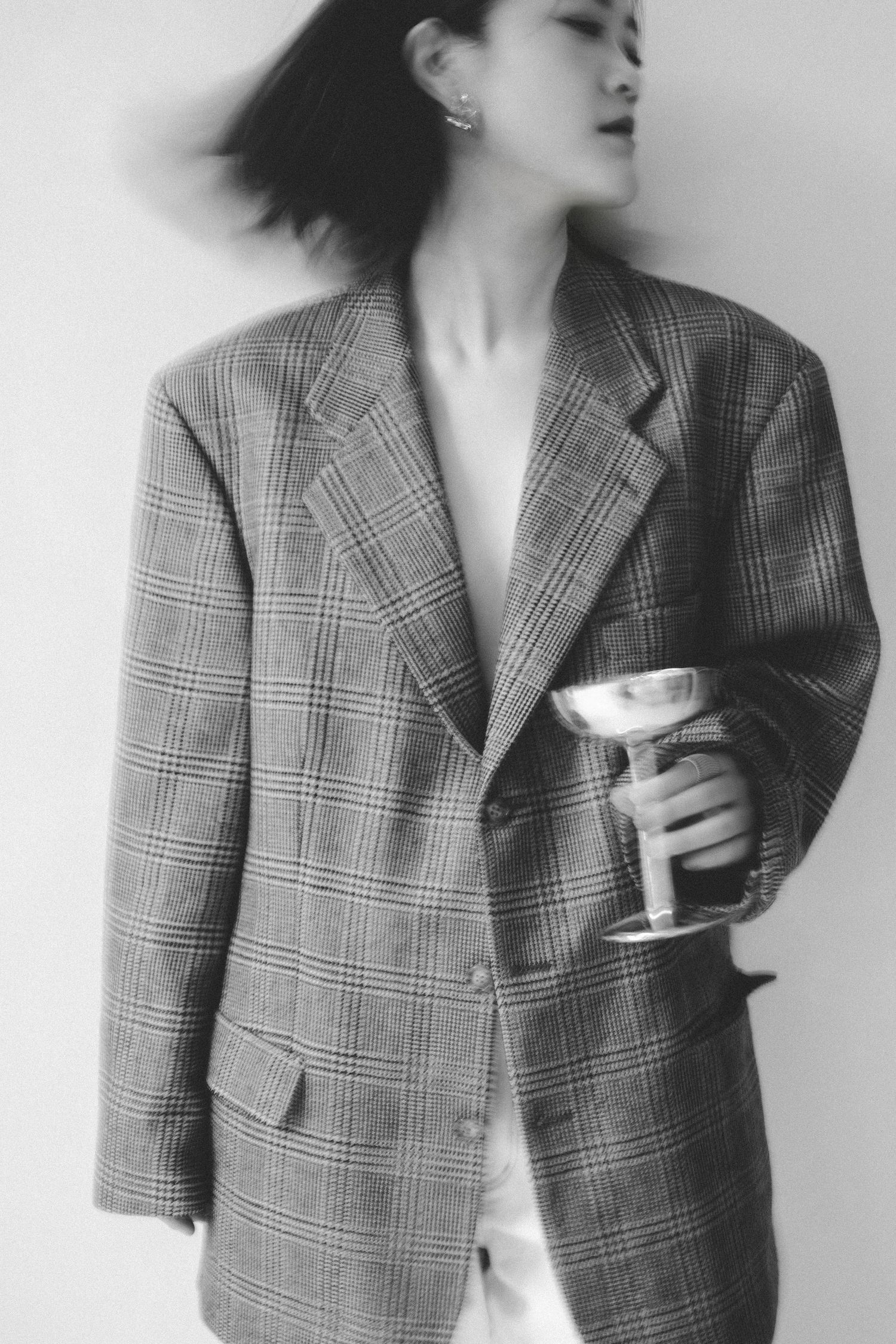 A figure in a plaid grey blazer jacket stands against a white background while holding a cup.