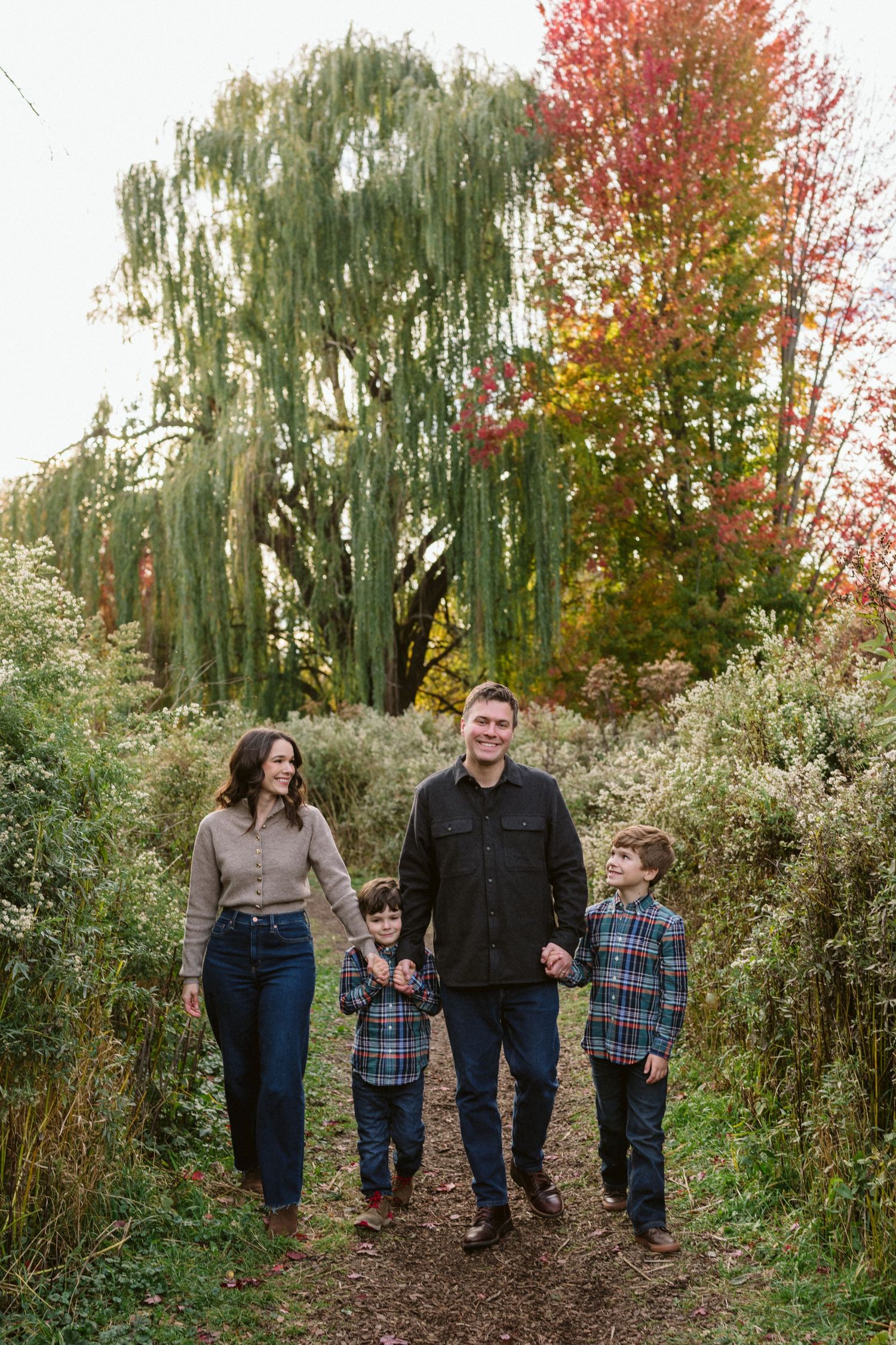 Family of four walking through autumn foliage path, parents holding hands with two boys in plaid shirts.