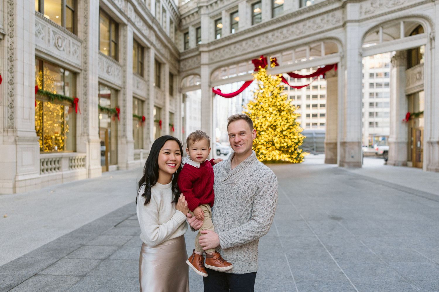Smiling couple holding a toddler in a red outfit in front of a lit golden Christmas tree and festive holiday decorations on a city street.