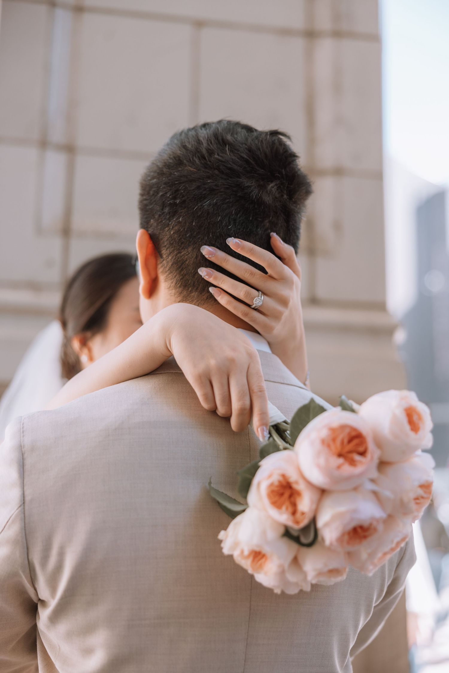 A romantic embrace between newlyweds, with a peach rose bouquet visible against a light gray suit jacket.
