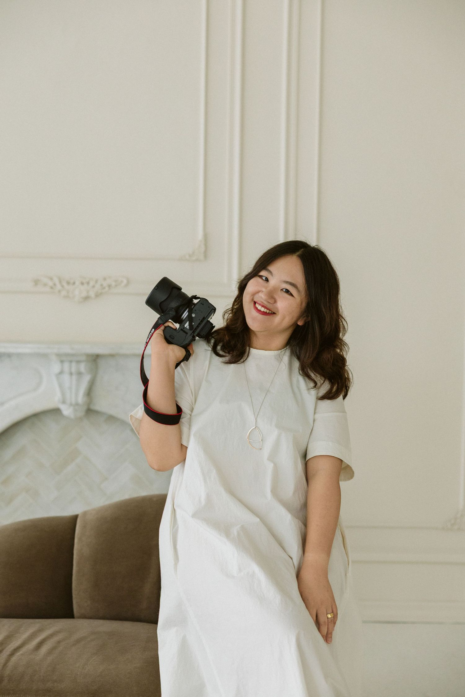 Smiling Asian woman in white dress holding a camera, posing near a sofa in a bright, elegant room.
