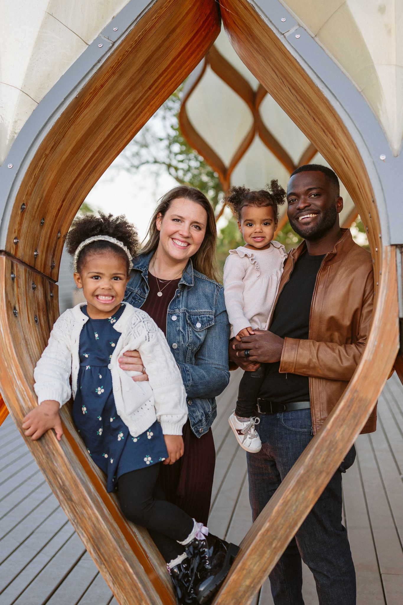 Happy interracial family of four posing inside a wooden arch structure outdoors, parents smiling with two young daughters.