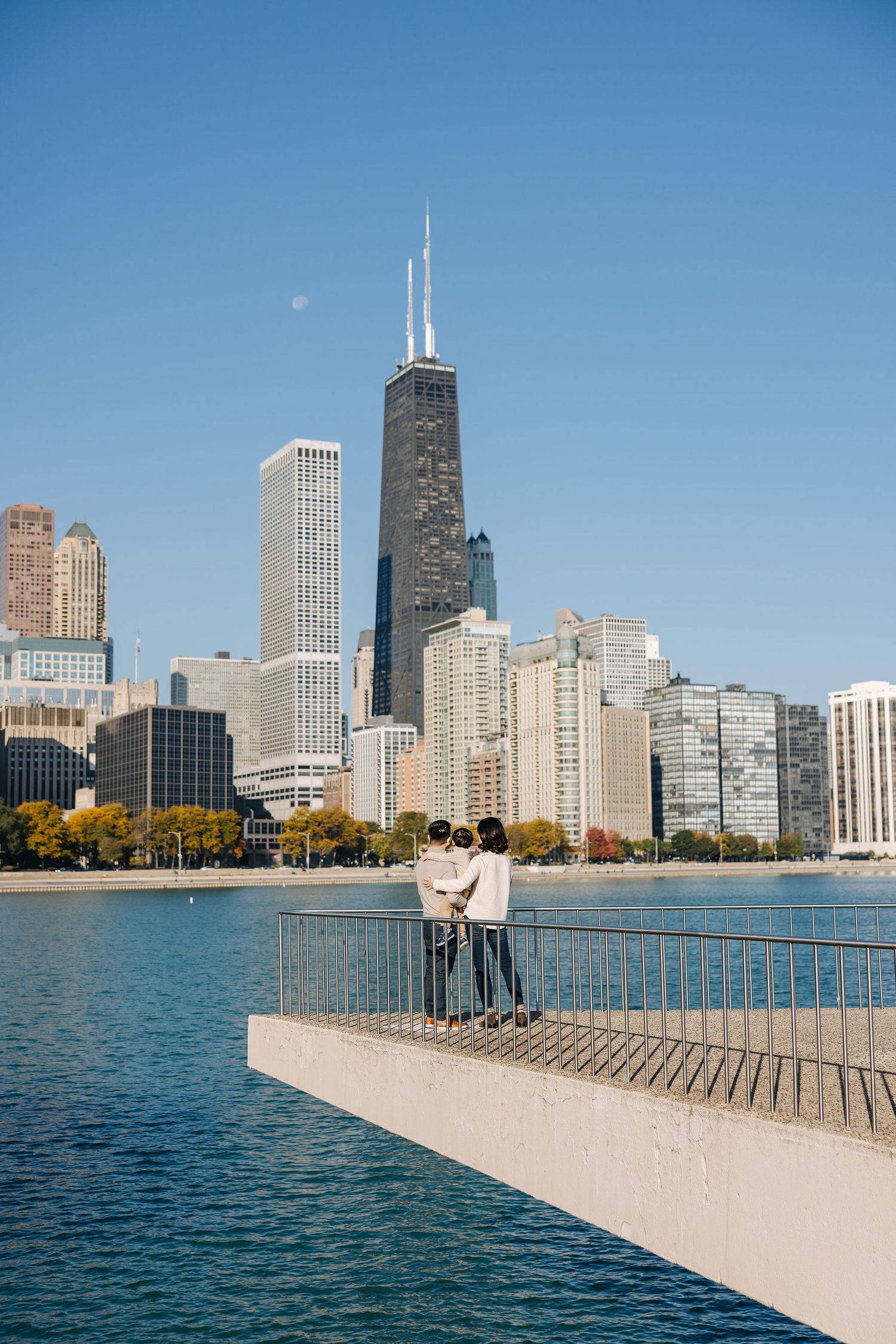 Couple standing on a pier overlooking Lake Michigan with the Chicago skyline and John Hancock Center on a clear day.