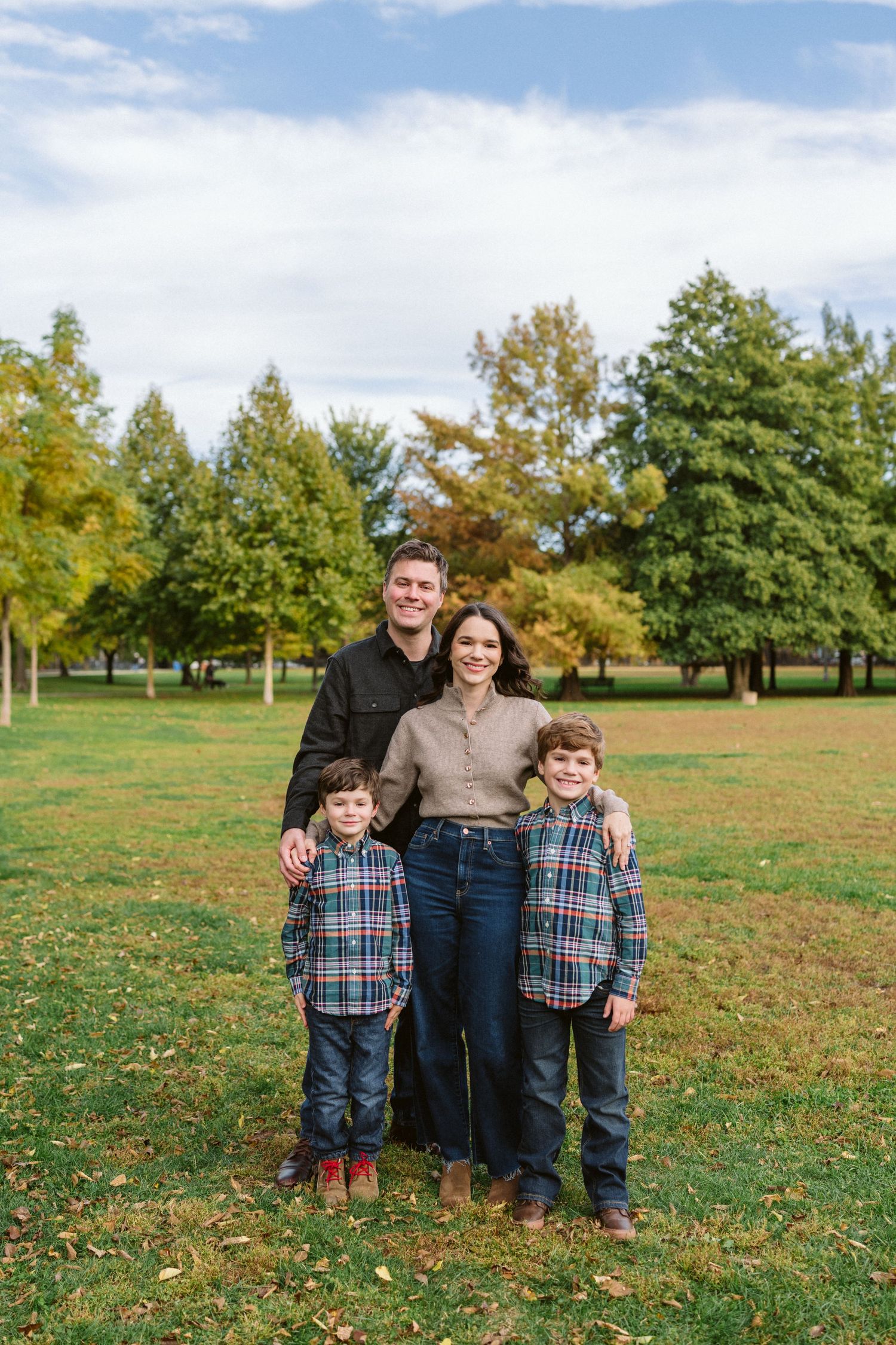 Family of four posing in a green park during autumn, parents and two boys in plaid shirts smiling outdoors.
