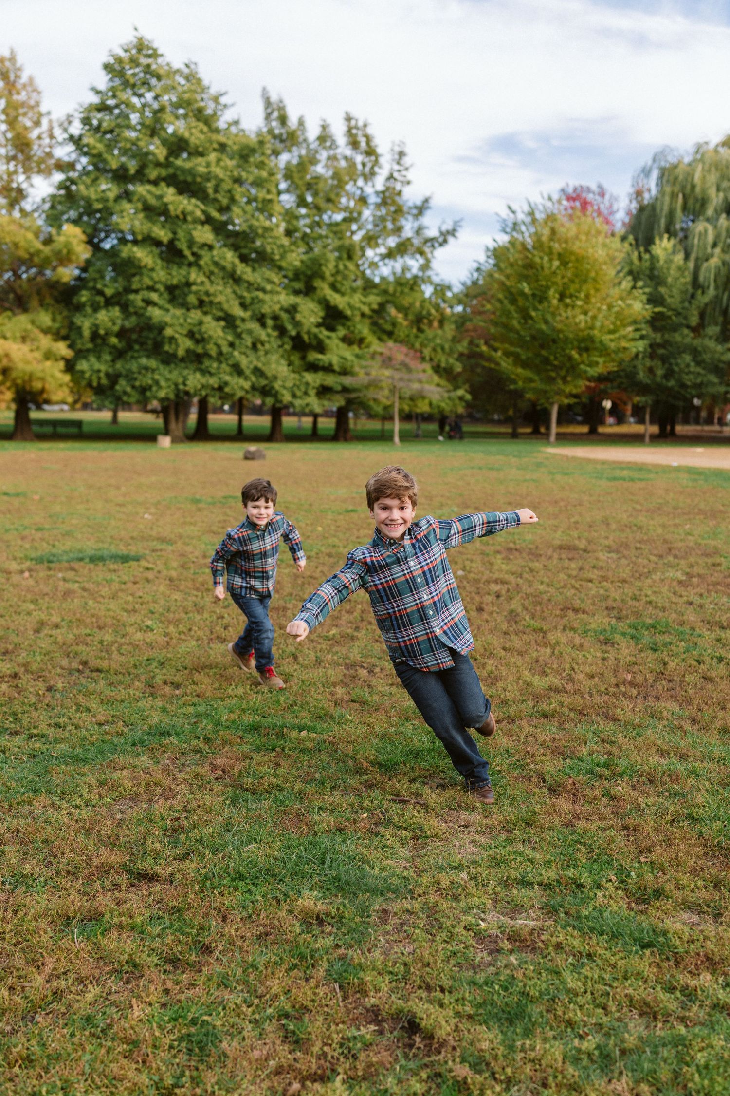 Two boys in plaid shirts running and playing in a green park with trees in the background.