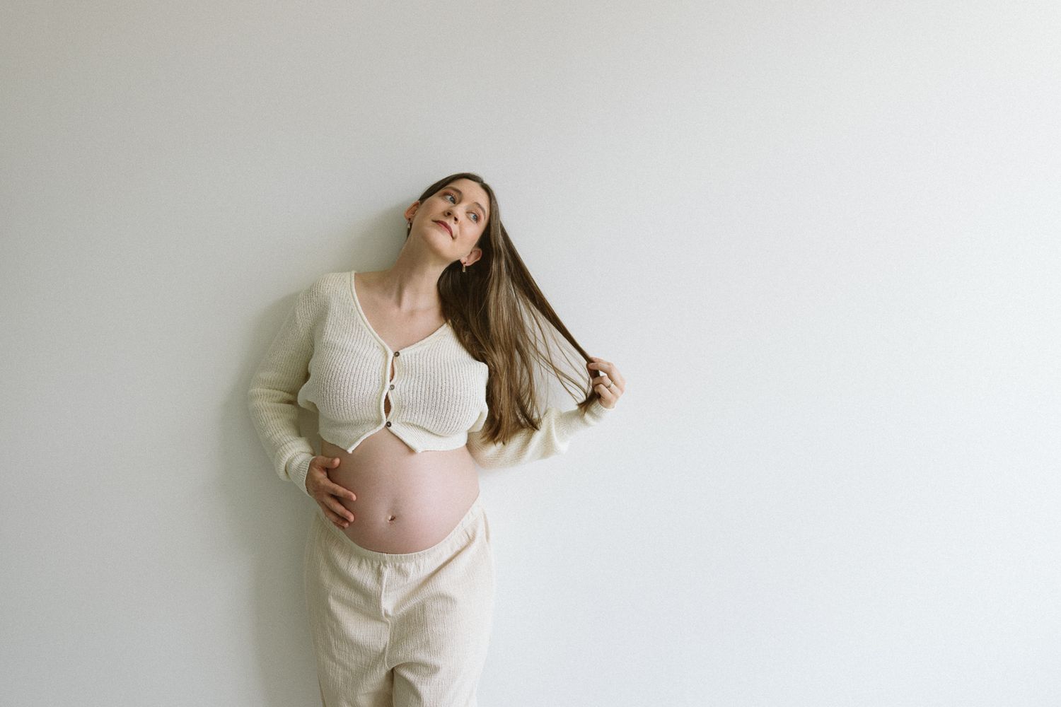 An expectant mother in white clothing caresses her baby bump against a plain wall.