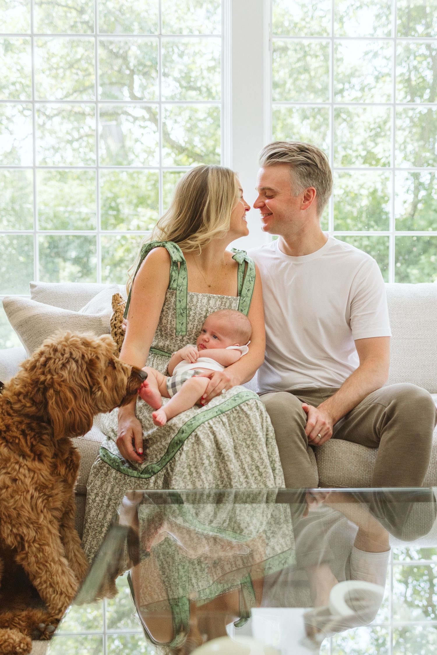 Family relaxing on couch with curly-haired goldendoodle dog and newborn baby in bright sunroom setting.