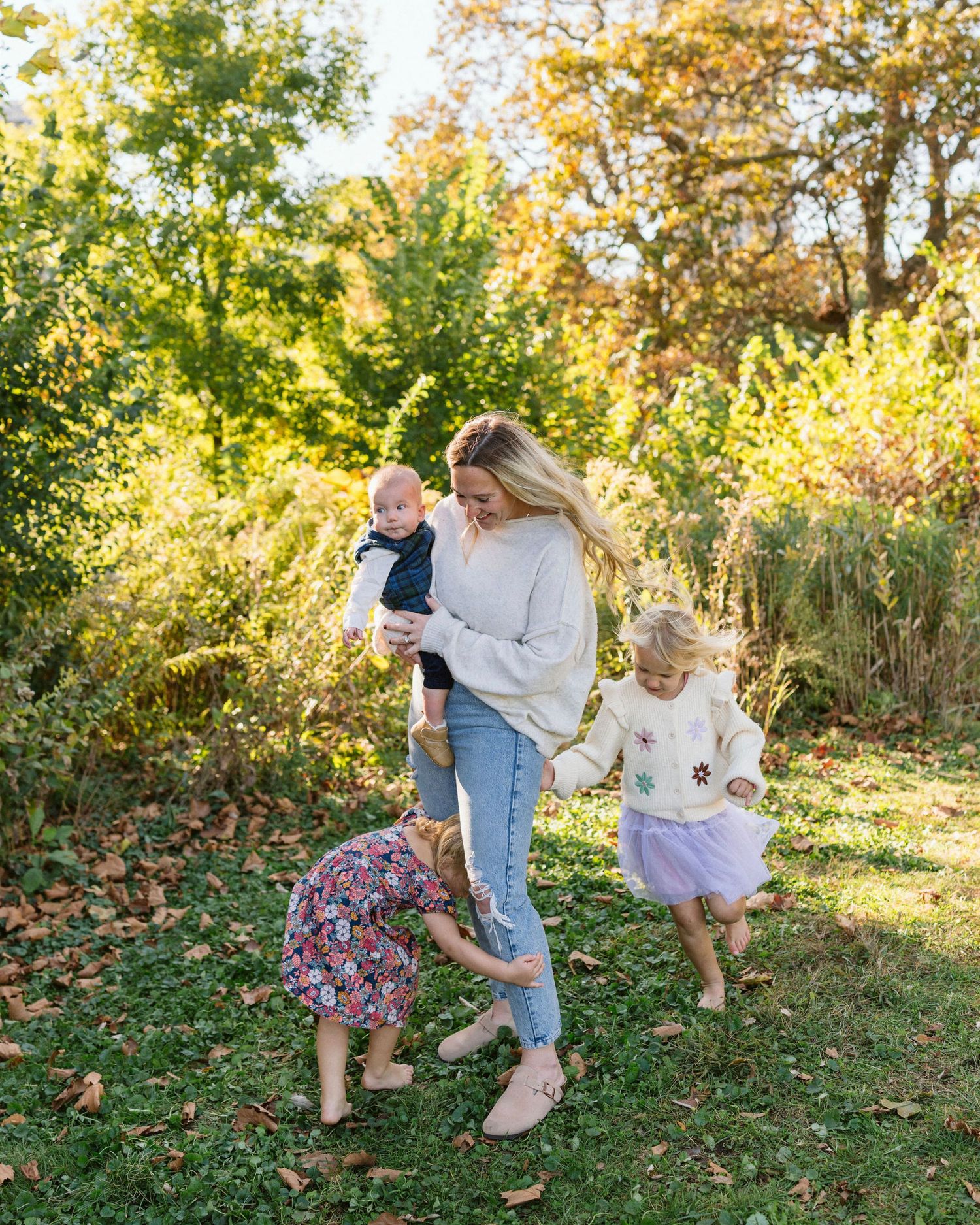 A woman holds a baby while playing outdoors with two young girls in a lush green park during autumn.