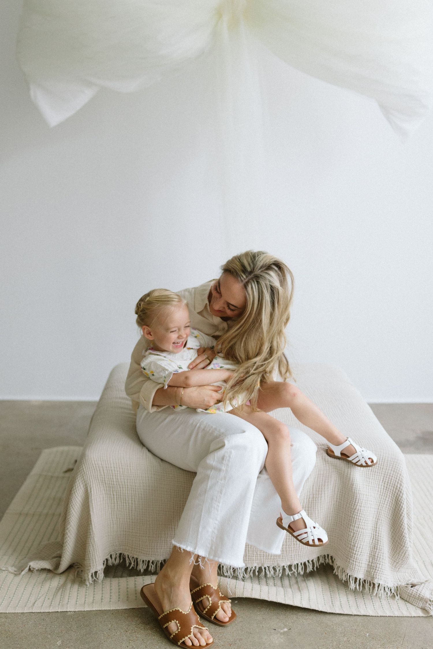 Two people in white dresses sit together on a cream-colored blanket in a bright studio setting.