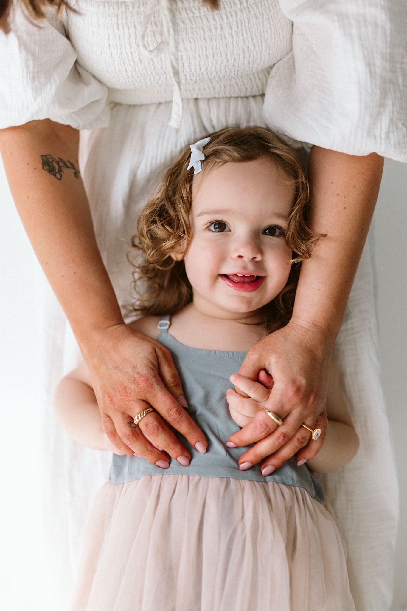 A small child in a blue and pink tulle dress stands between adult hands wearing a white top with visible tattoo.