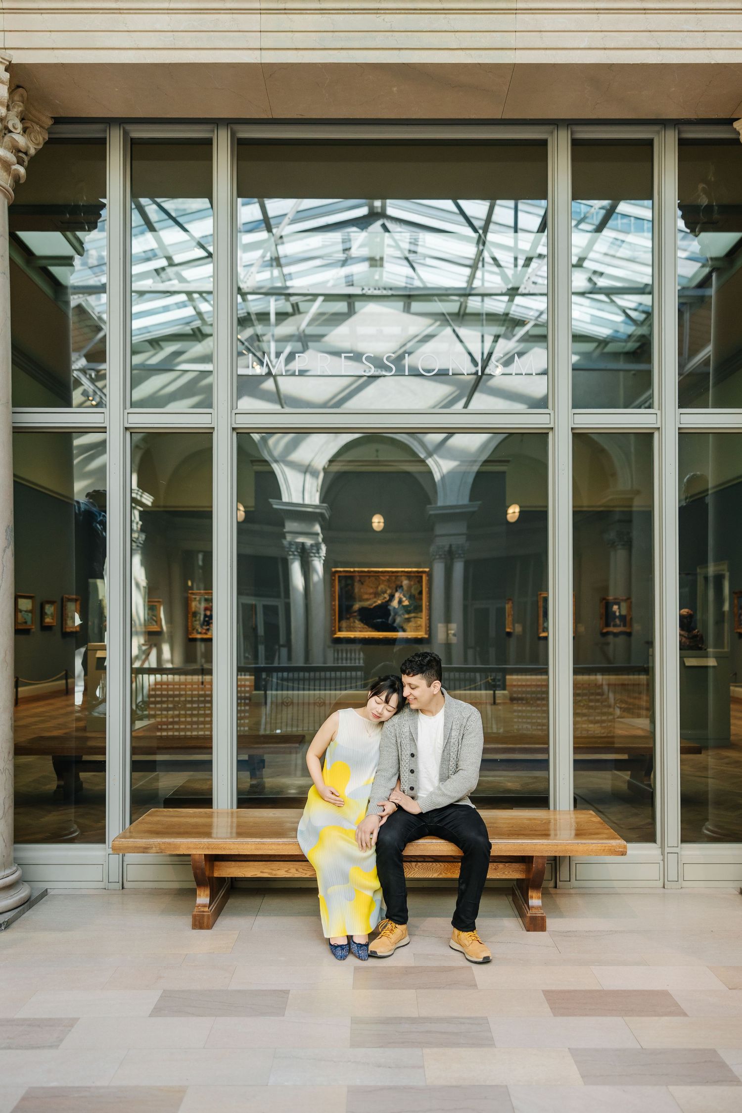A couple sits together on a wooden bench inside a building with glass windows and classic architectural details.