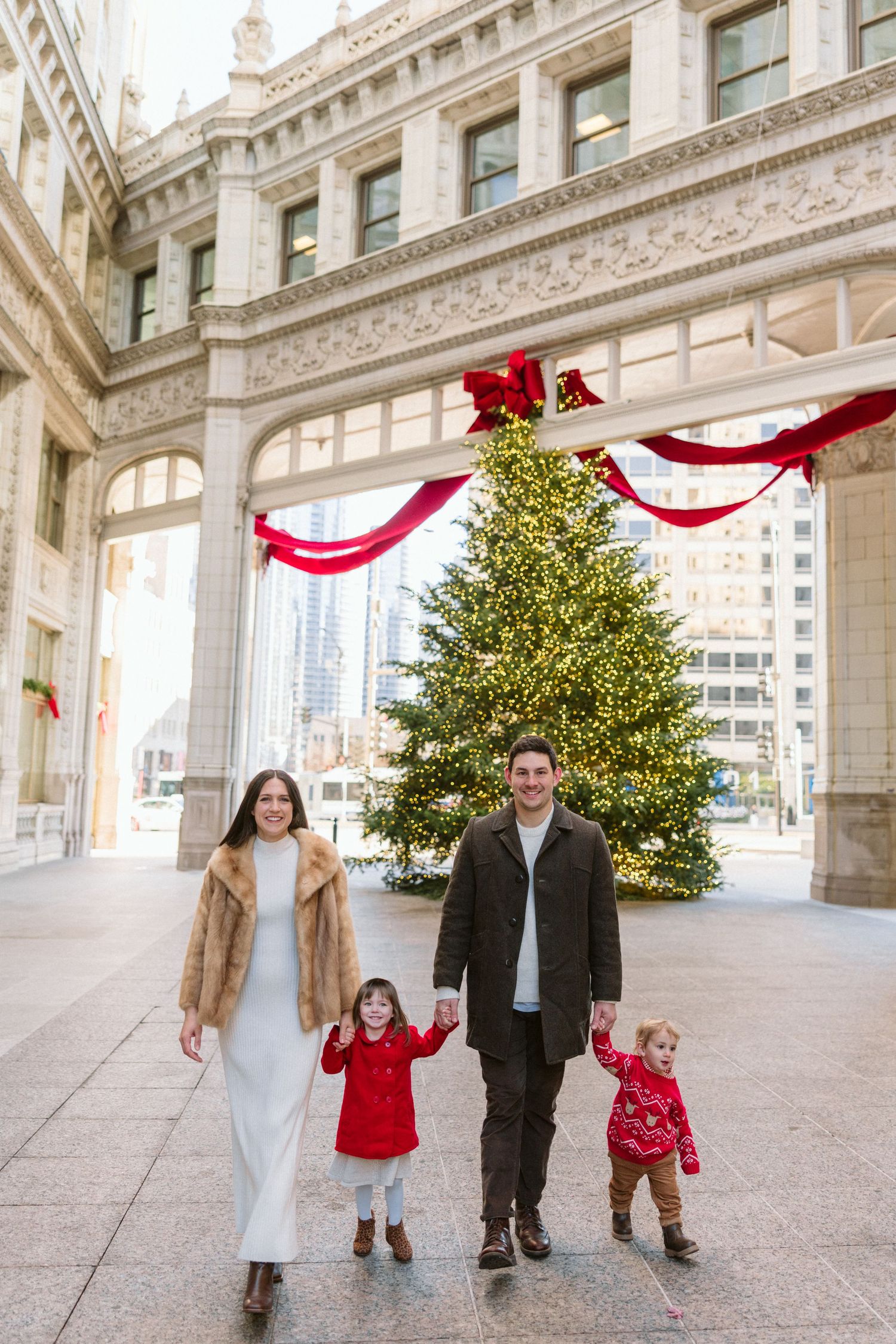 Family of four walking hand in hand beneath a decorated archway with a lit Christmas tree and red ribbons in a city plaza.