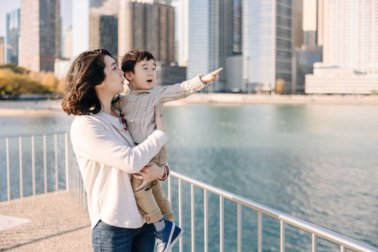 Mother holds toddler son while pointing at city skyline view from waterfront promenade on sunny day.