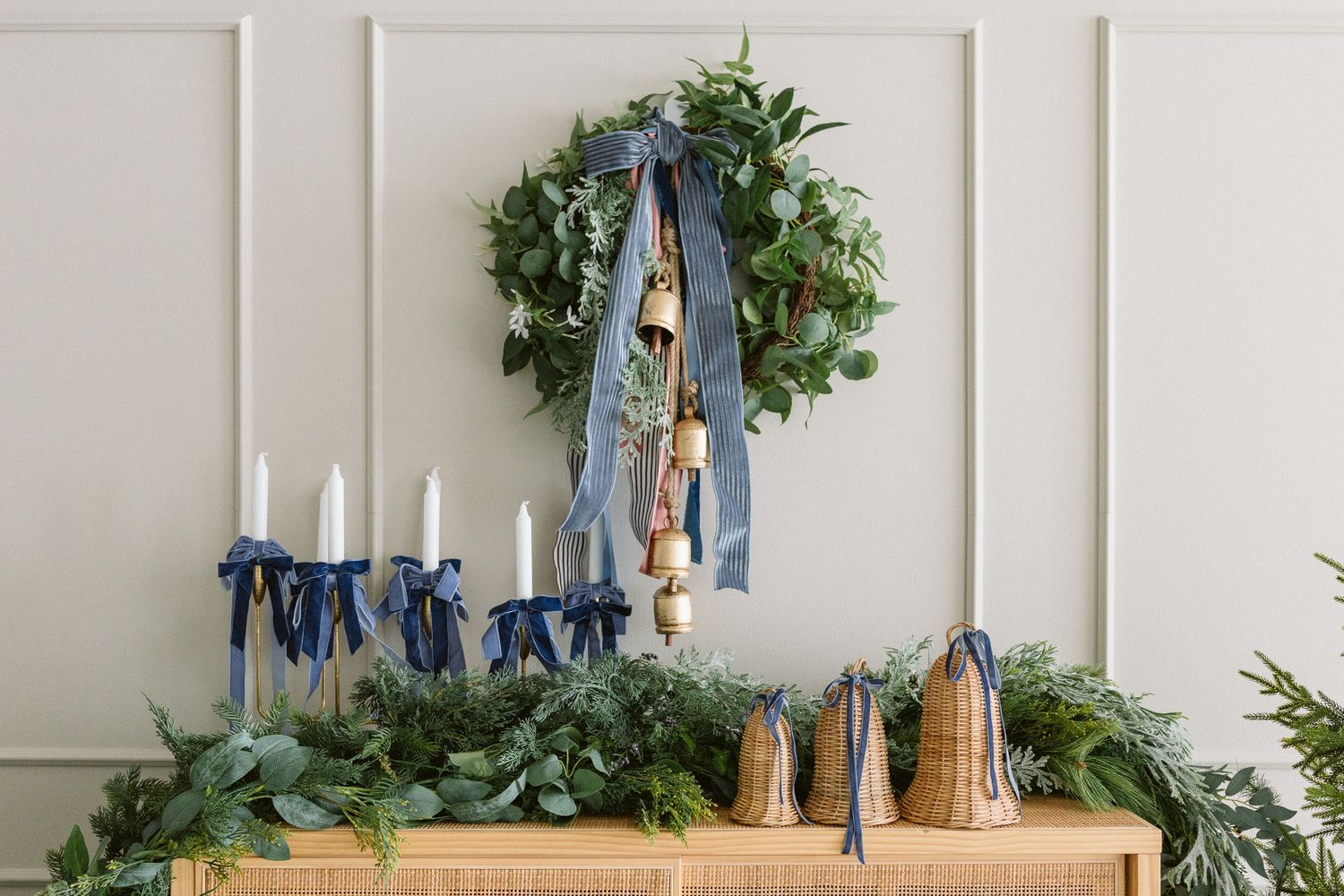 Evergreen wreath with blue velvet ribbon and brass bells hangs above a rattan mantel styled with greenery, candles, and bell ornaments.