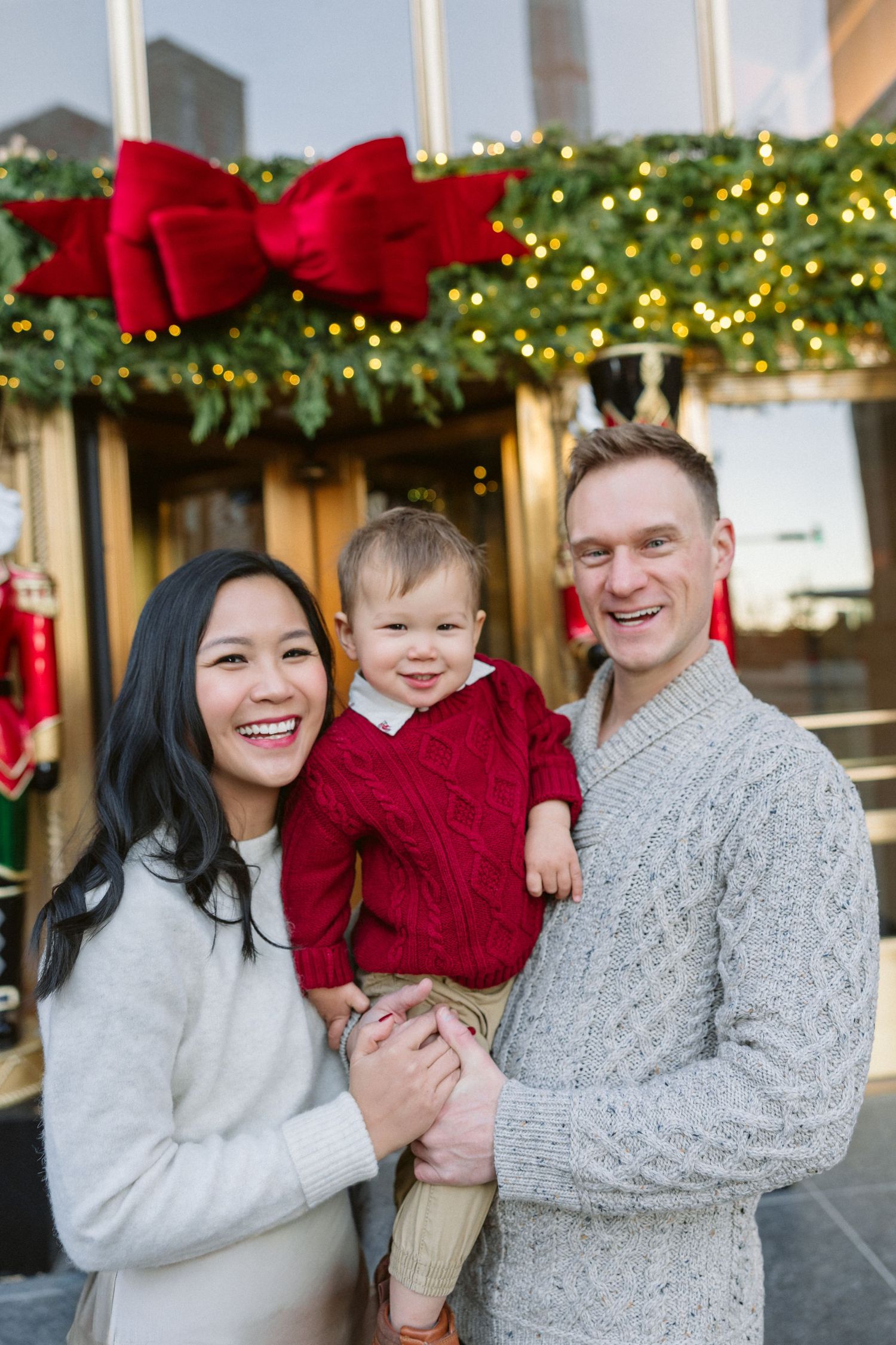 Smiling couple holding a toddler in a red sweater outside a building decorated with a holiday garland, lights, and a large red bow.