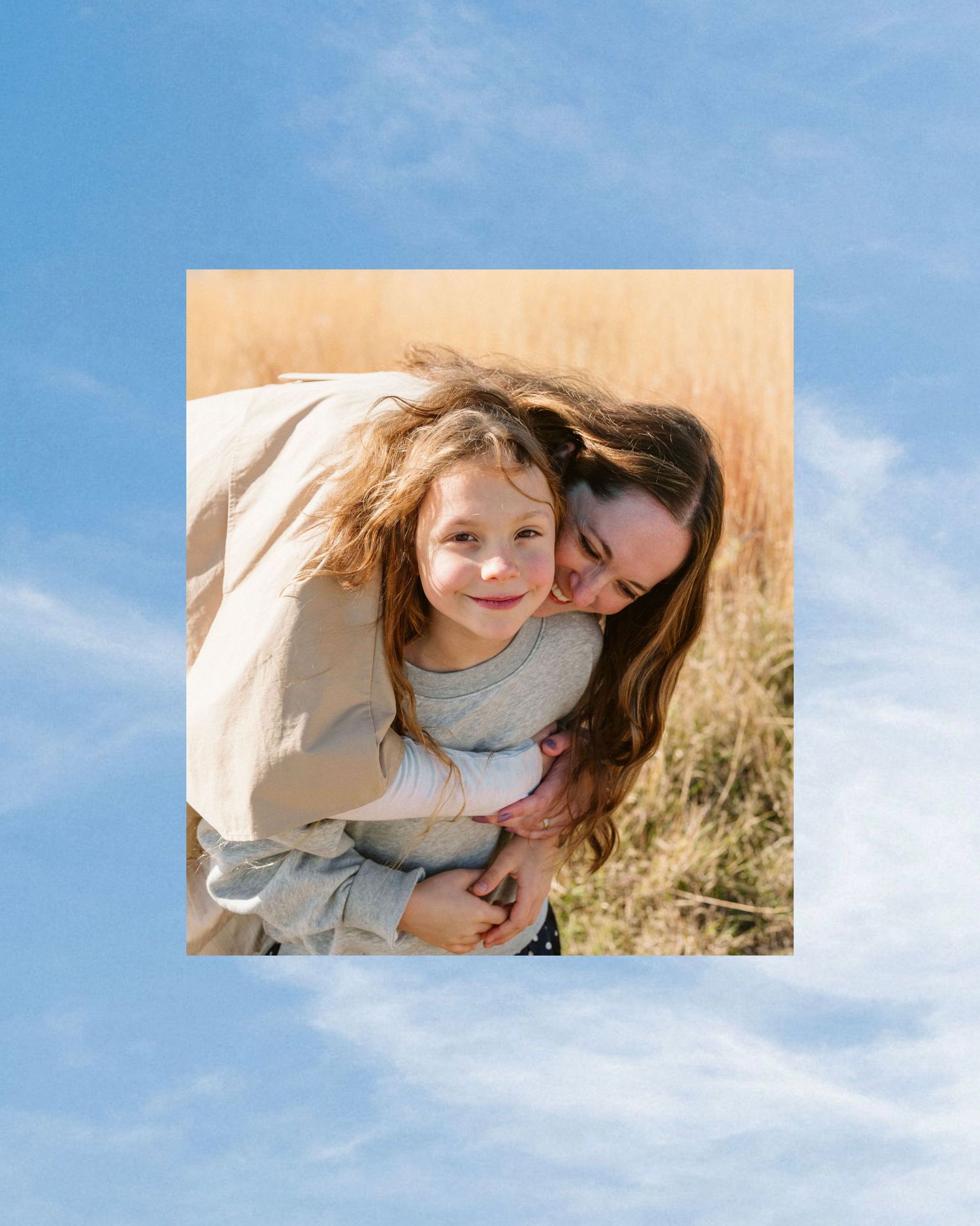 A mother hugging her young daughter in a golden field under a bright blue sky.
