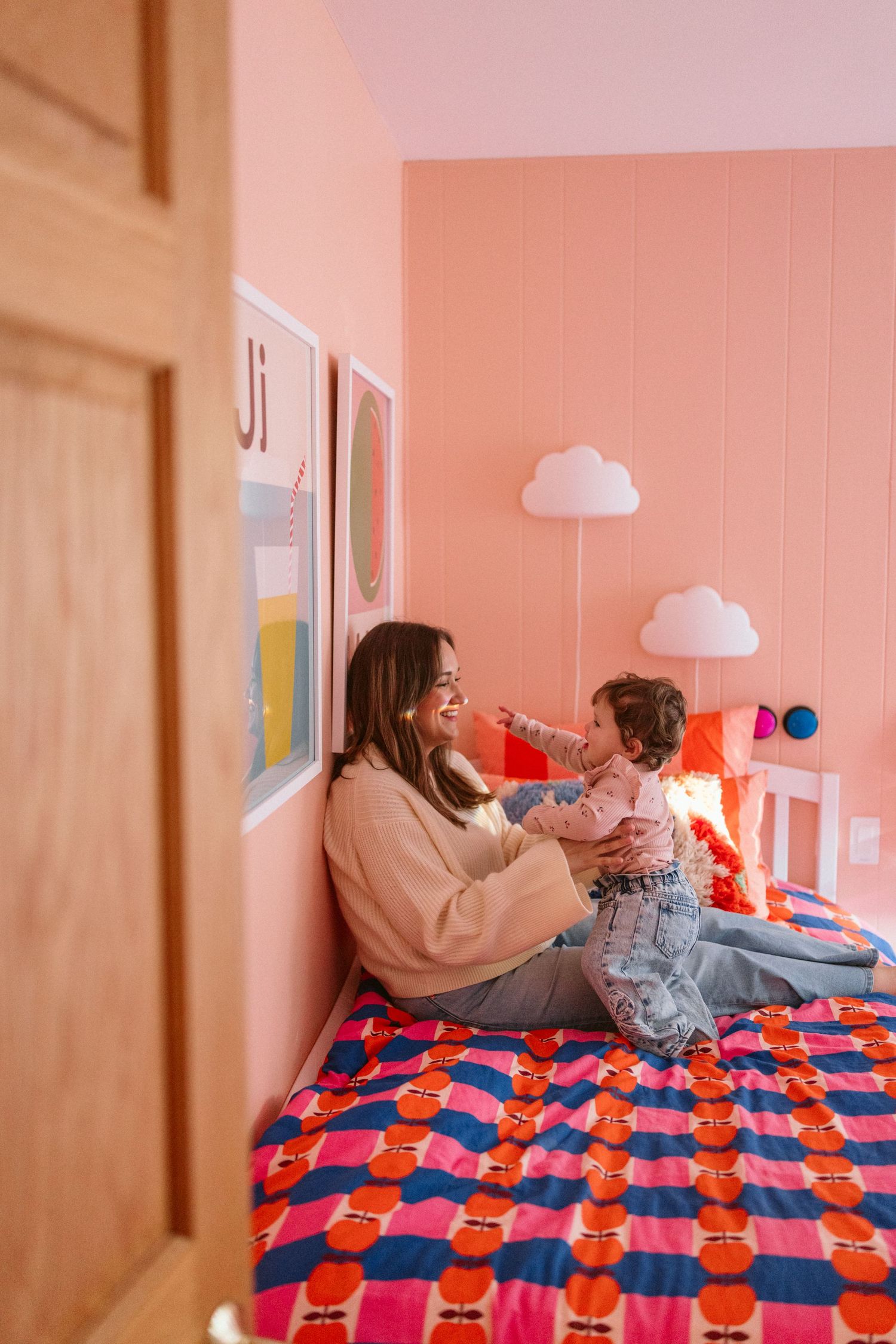 A mother and toddler playing together on a colorful bed in a pink bedroom with cloud wall decor.