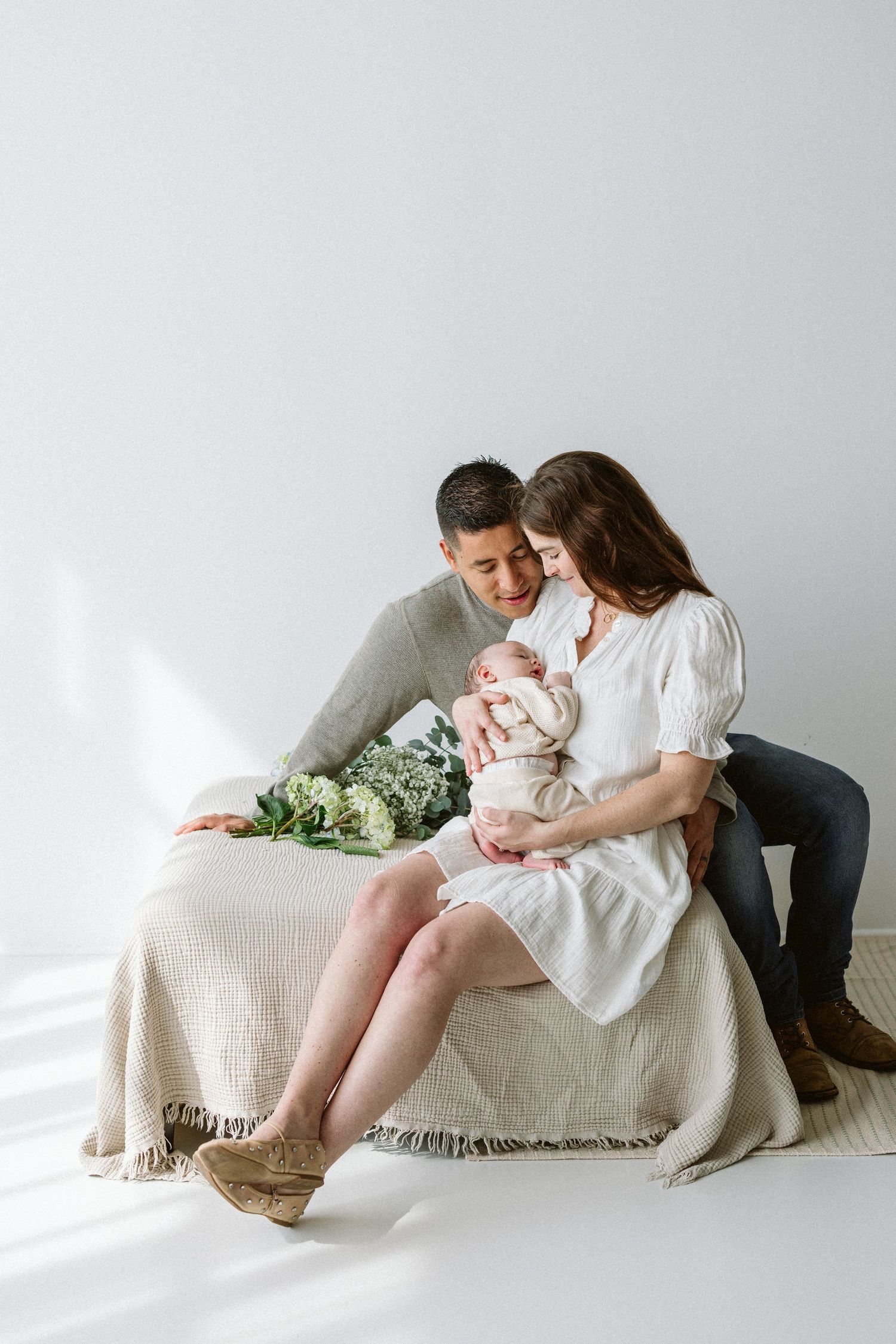 Family snuggles together on white couch with soft beige blanket in minimalist studio setting.