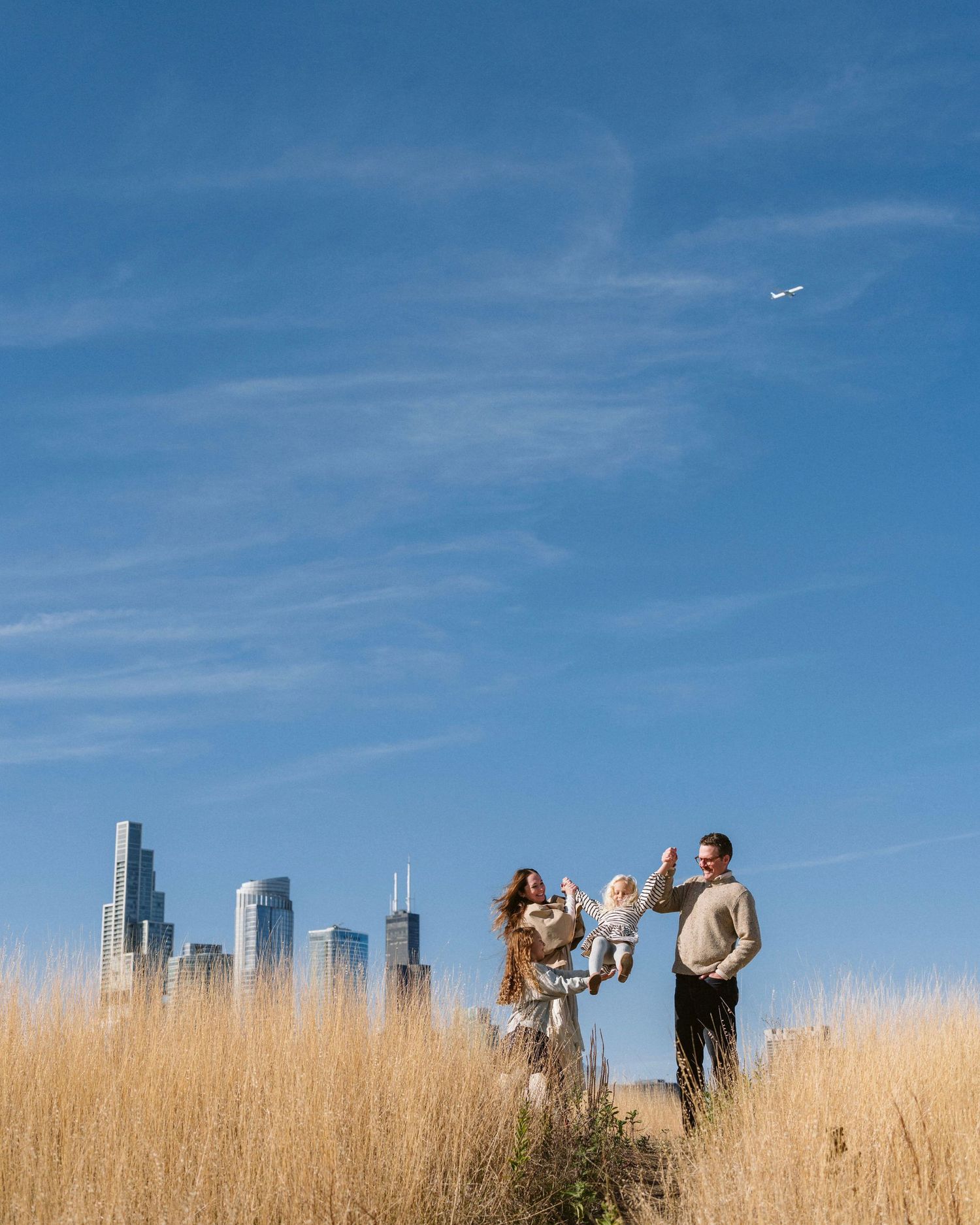 Family of three playing in golden grass fields with Chicago skyline and blue sky backdrop on sunny day.