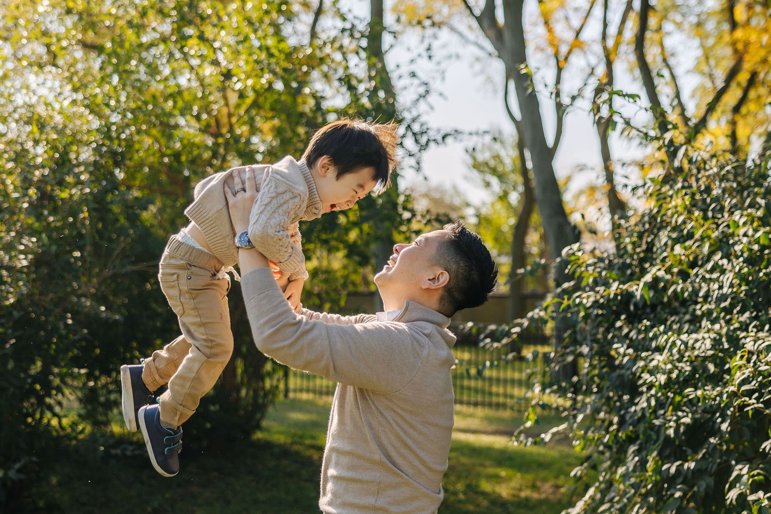 Father joyfully lifting young child in sunny autumn park, both smiling, surrounded by green and golden foliage.