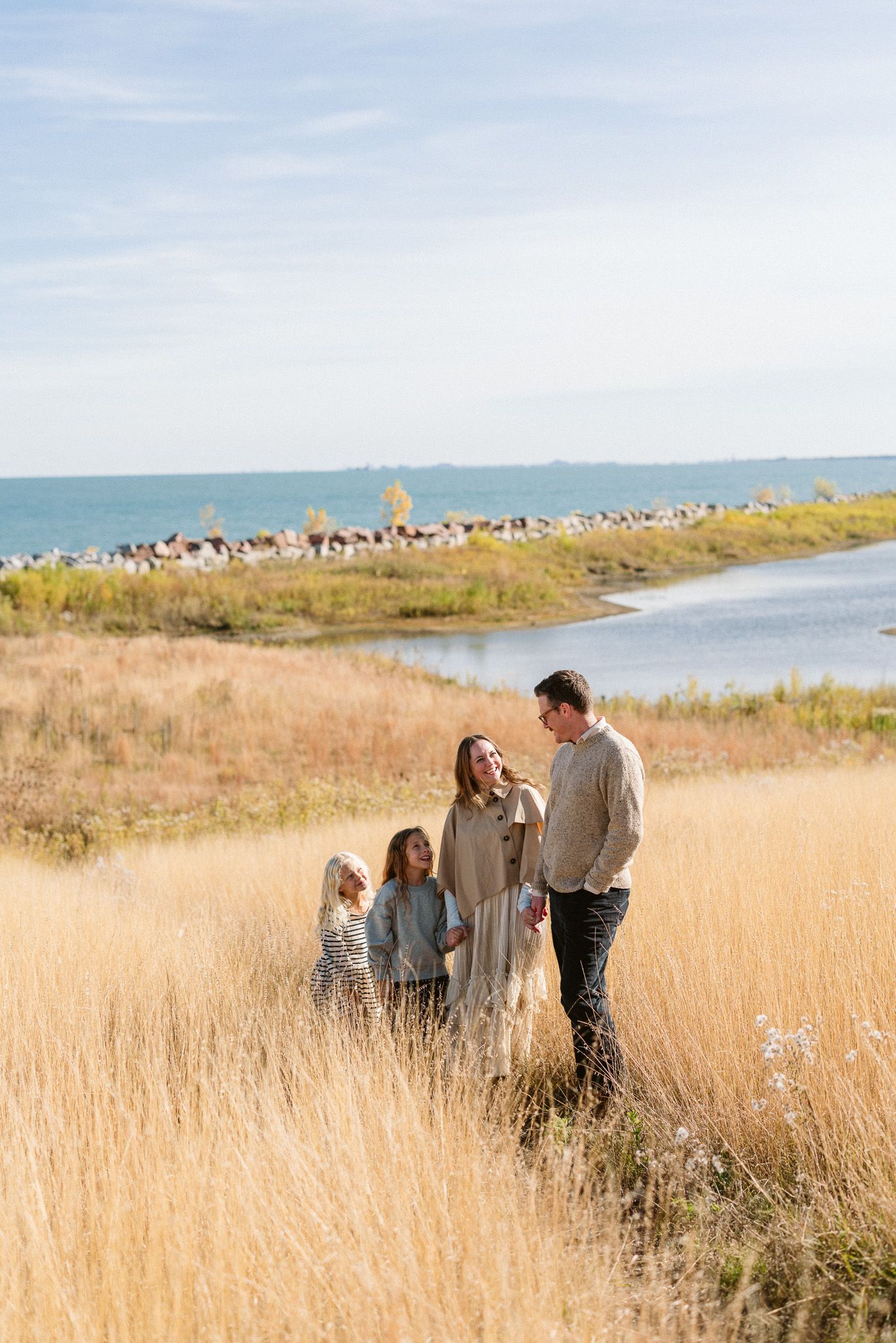 Family of four standing in golden tall grass near a serene lake on a sunny day in casual neutral outfits.