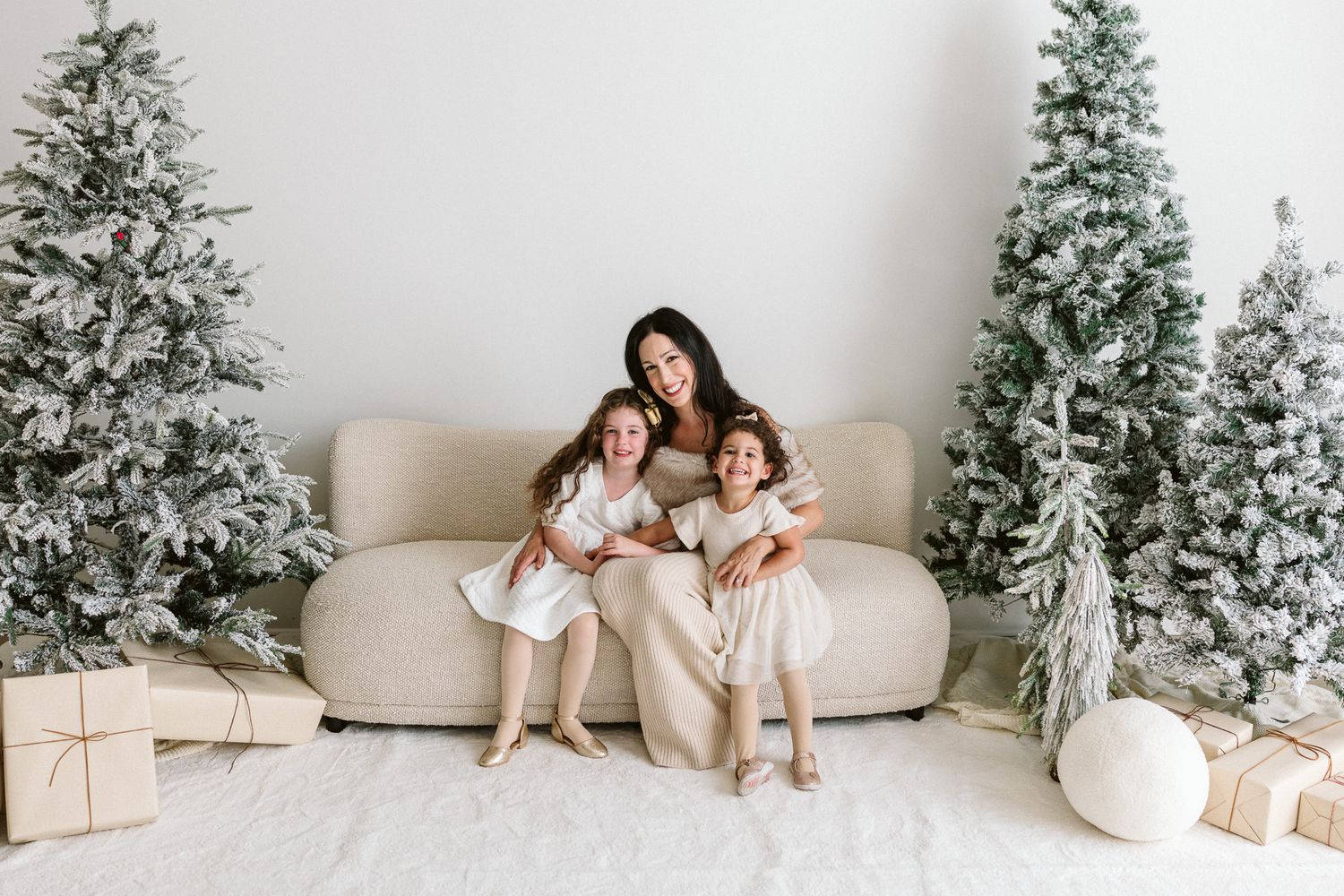 Mother and two young daughters in cream outfits cuddle on a beige sofa surrounded by flocked Christmas trees and wrapped gifts.