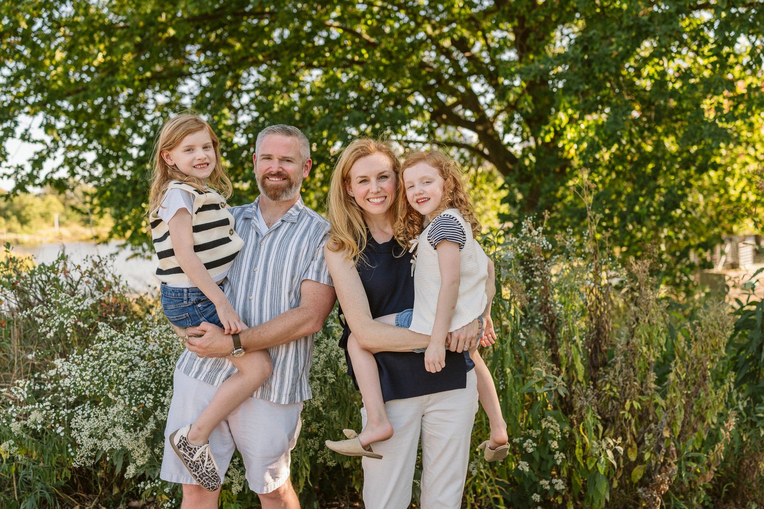 Family of four posing outdoors among lush green trees, parents holding two young red-haired girls, smiling warmly.