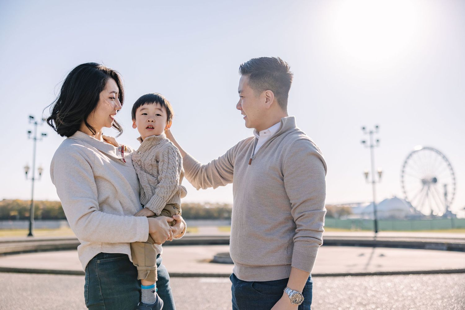 Asian family with mother holding toddler and father touching child's face outdoors near a Ferris wheel on a sunny day.
