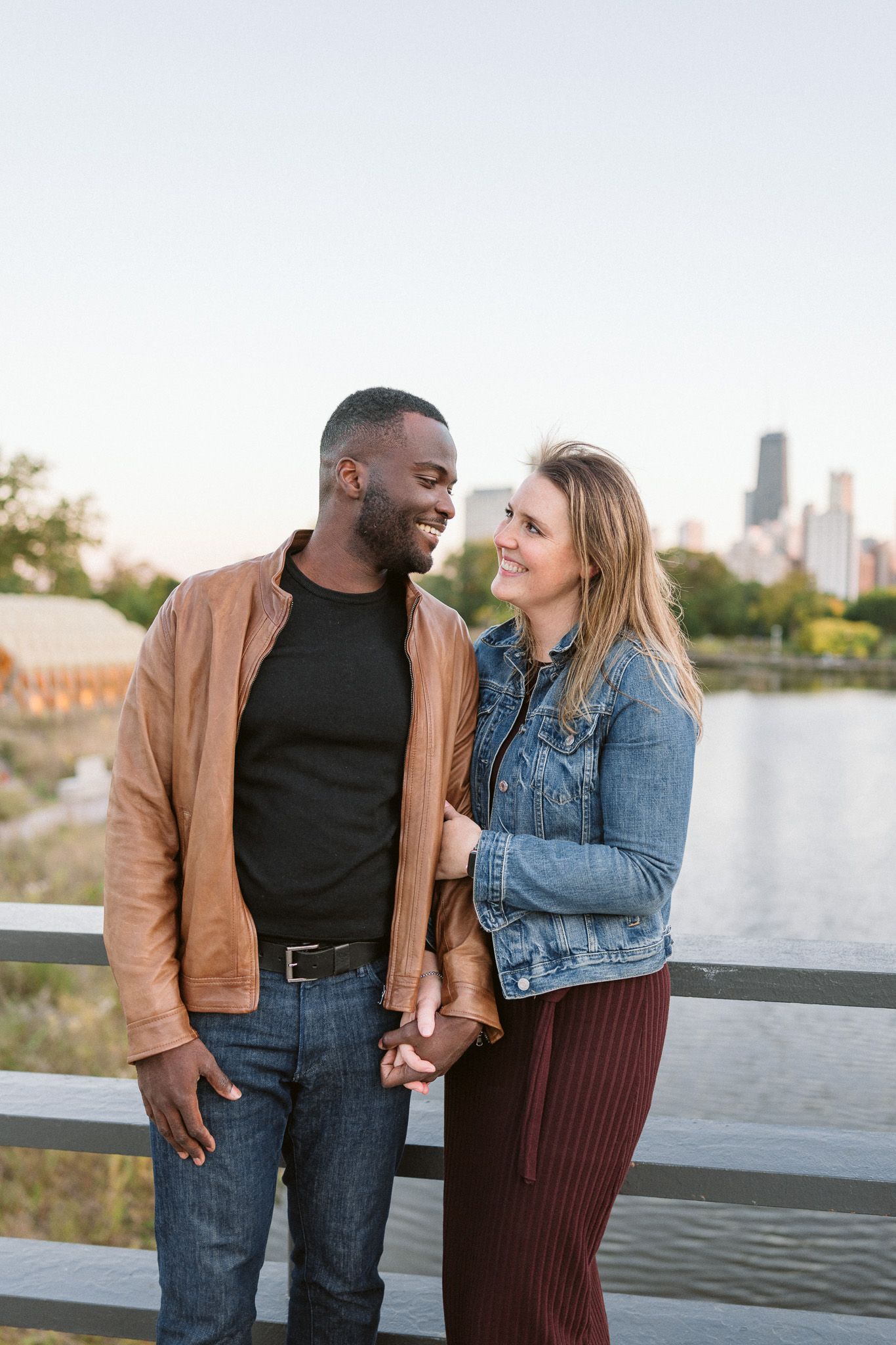 Interracial couple holding hands and smiling at each other on a bridge with a city skyline backdrop.