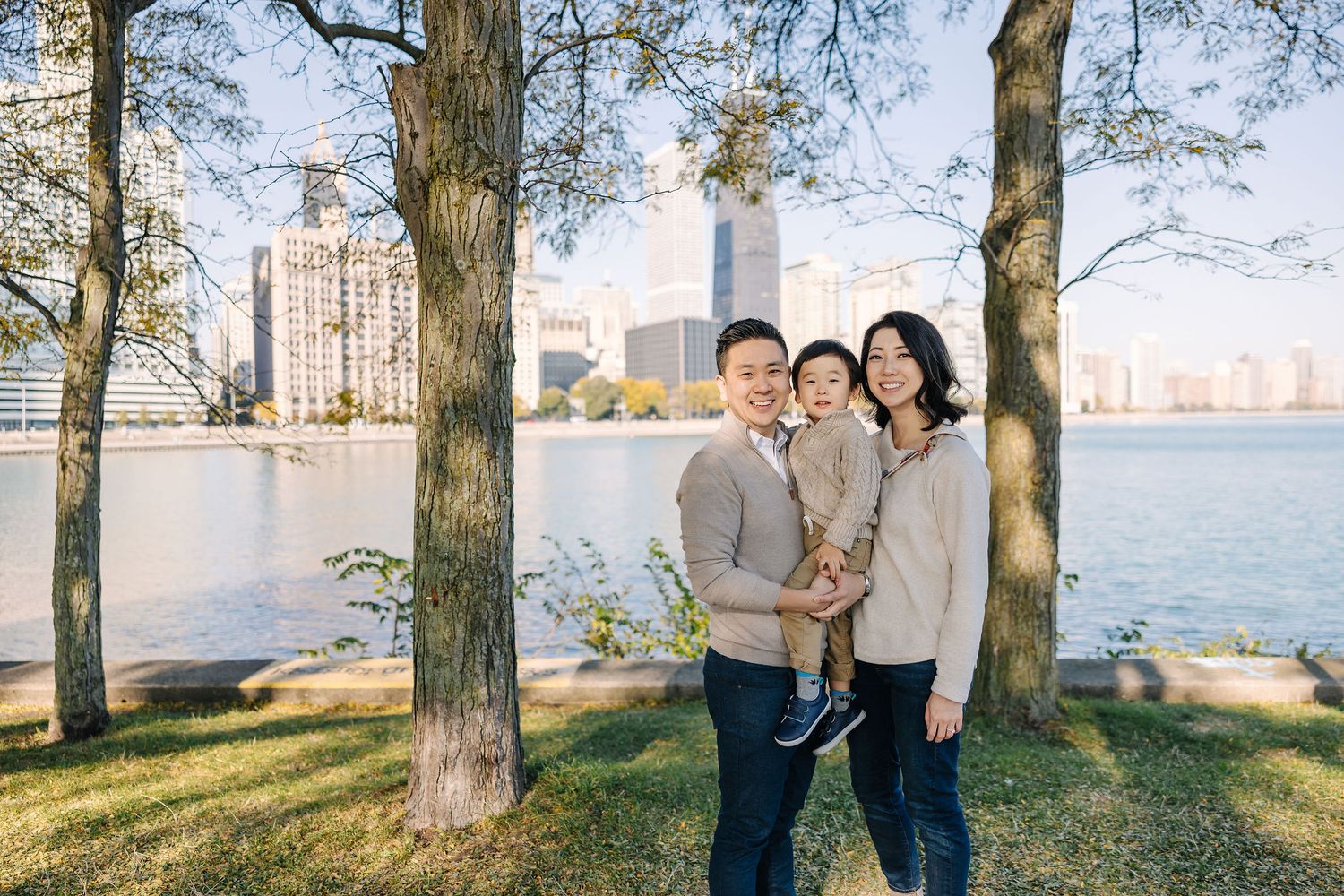 Asian family of three poses by lakeside with city skyline backdrop amid autumn trees on a sunny day.